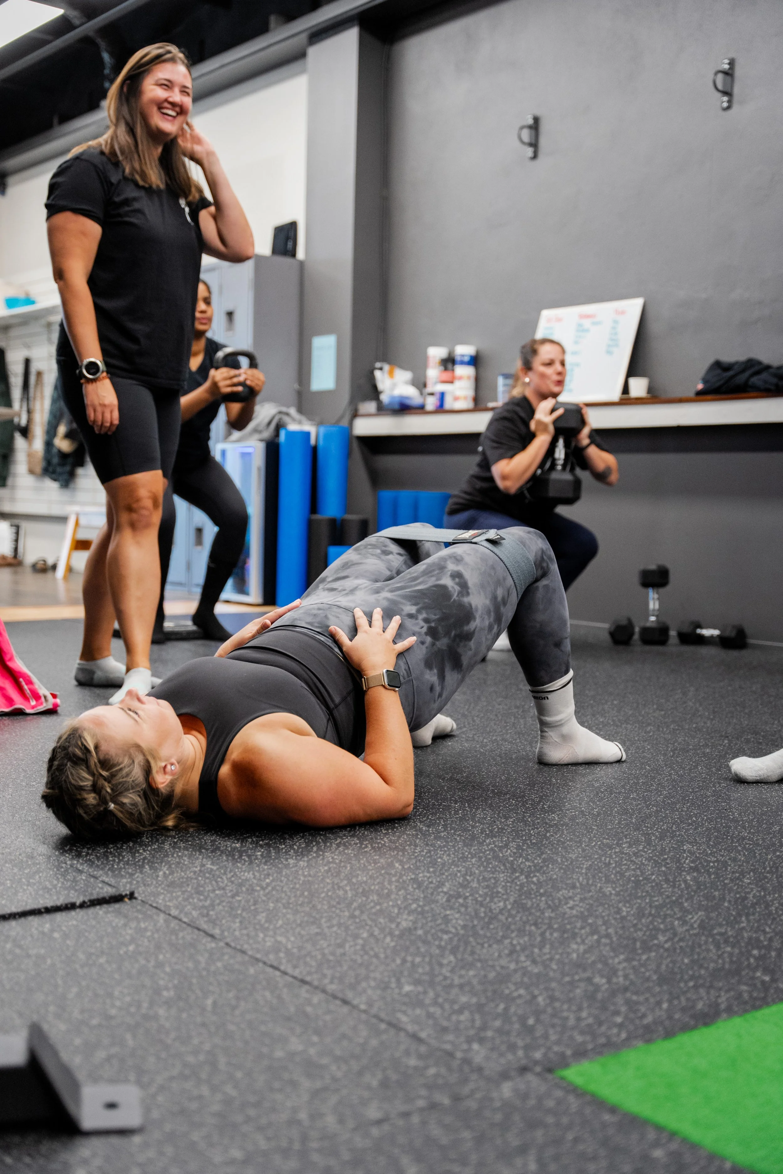 A woman lying on the floor doing a glute bridge exercise with her knees bent and hands on her abdomen, in a gym. Two women are standing nearby, one smiling and the other holding a coffee cup, and a woman in the background is taking photos. Fitness equipment is visible around them.