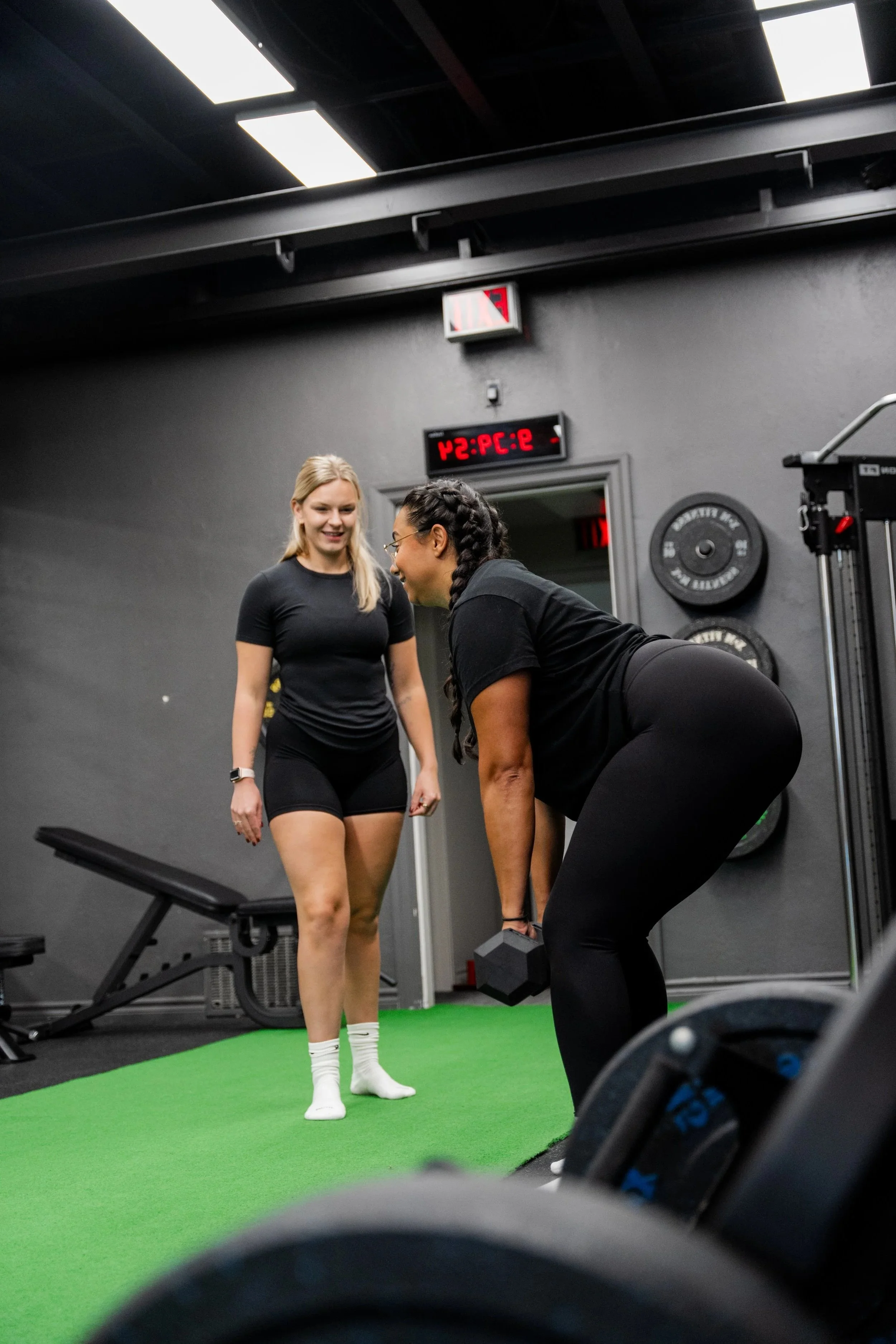 Two women exercising in a gym, one bending over lifting a dumbbell and the other standing nearby smiling.