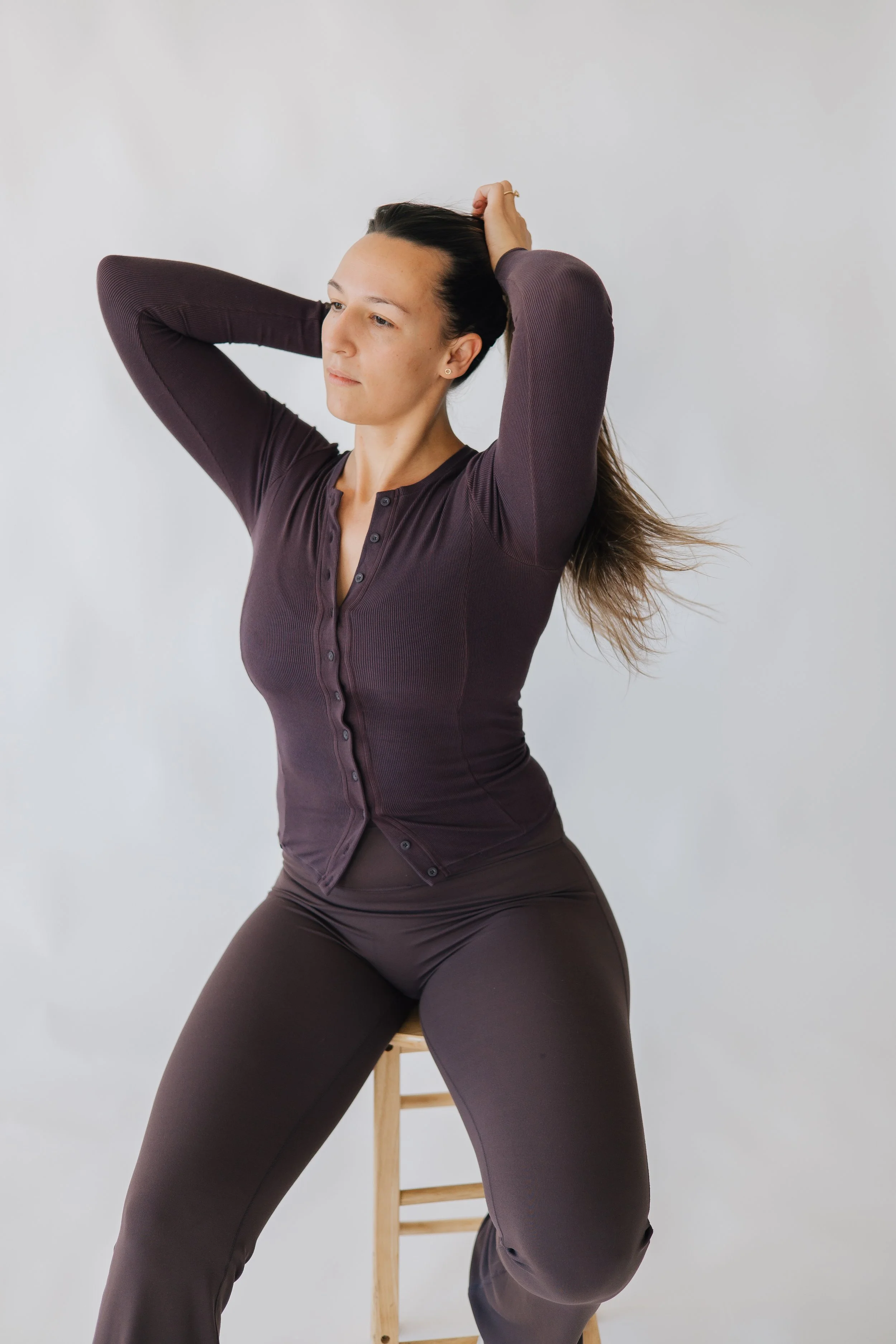 Woman in dark long-sleeve shirt and matching pants sitting on a wooden stool with her hands in her hair, looking to the side against a plain white background.