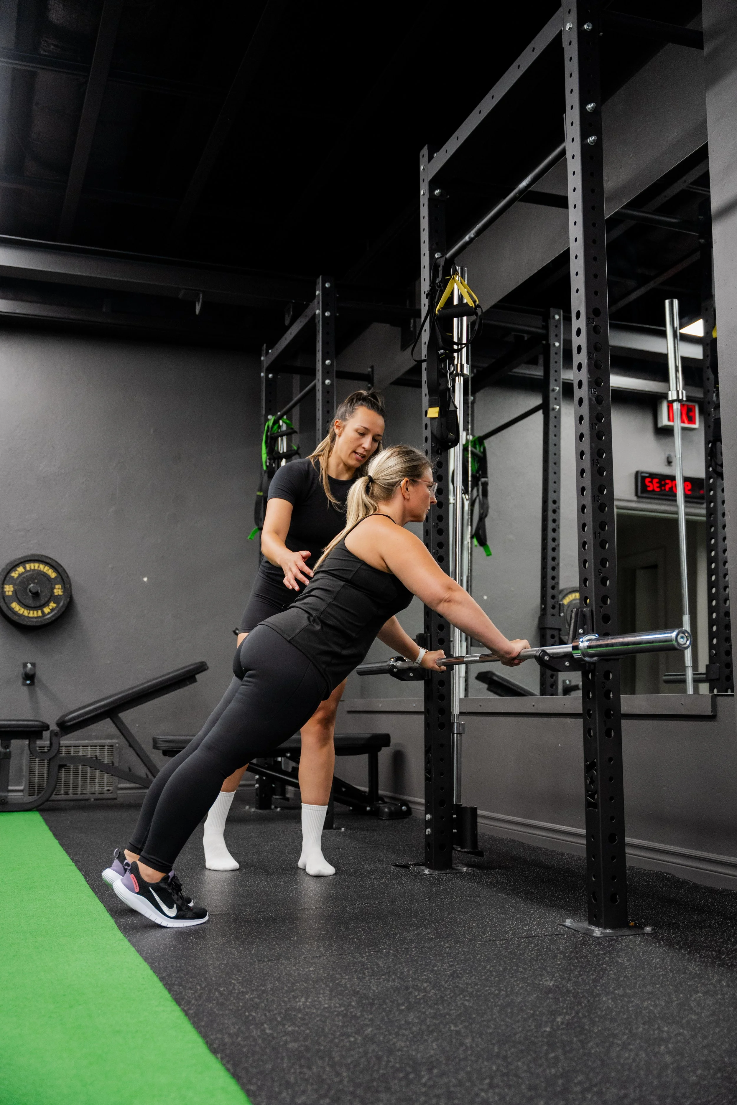 Woman performing a barbell exercise with supervision from another woman in a gym with black walls, gym equipment, and a green turf area.