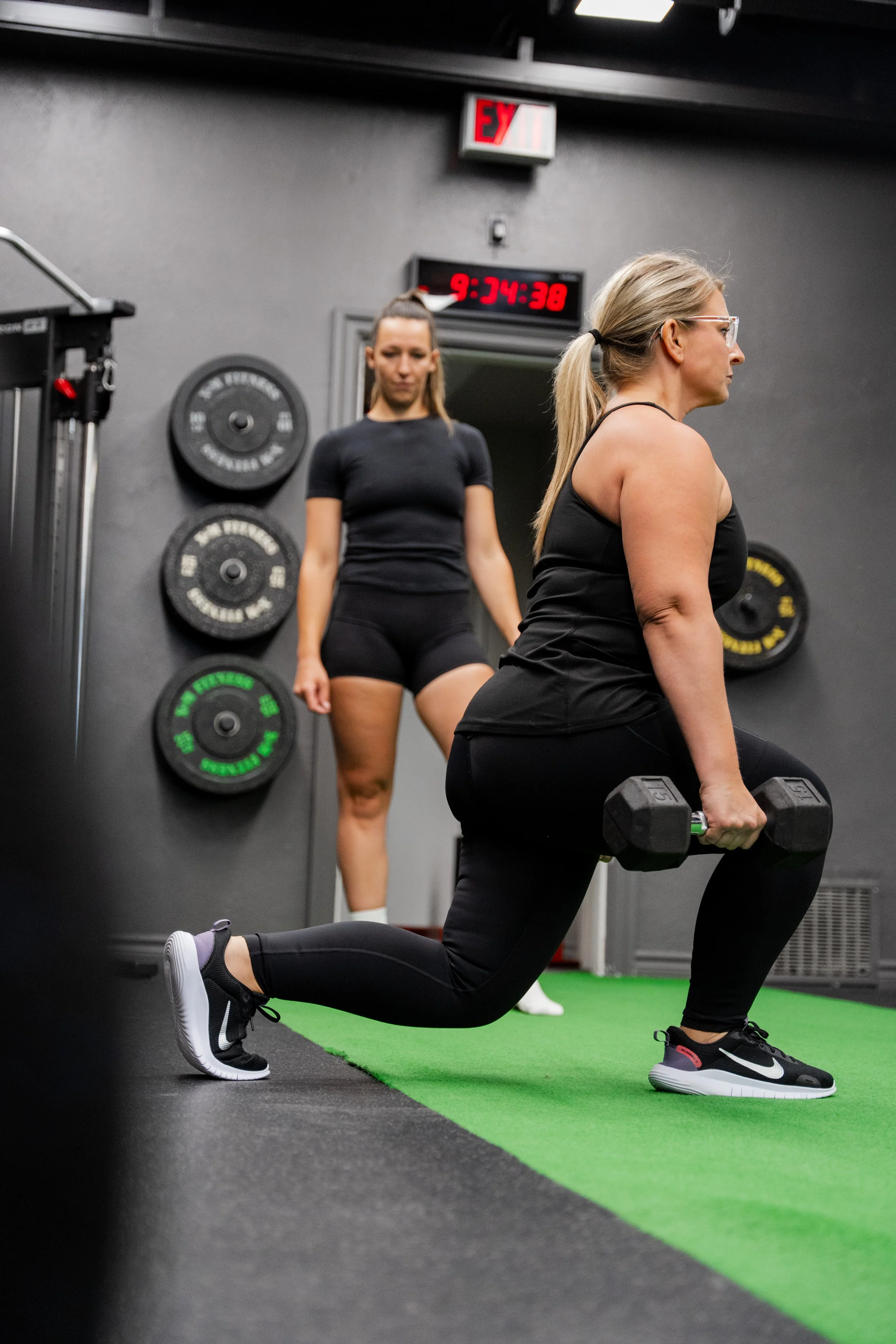 A woman in black workout clothes and sports shoes performs a lunge exercise holding dumbbells in a gym, with another woman watching in the background.