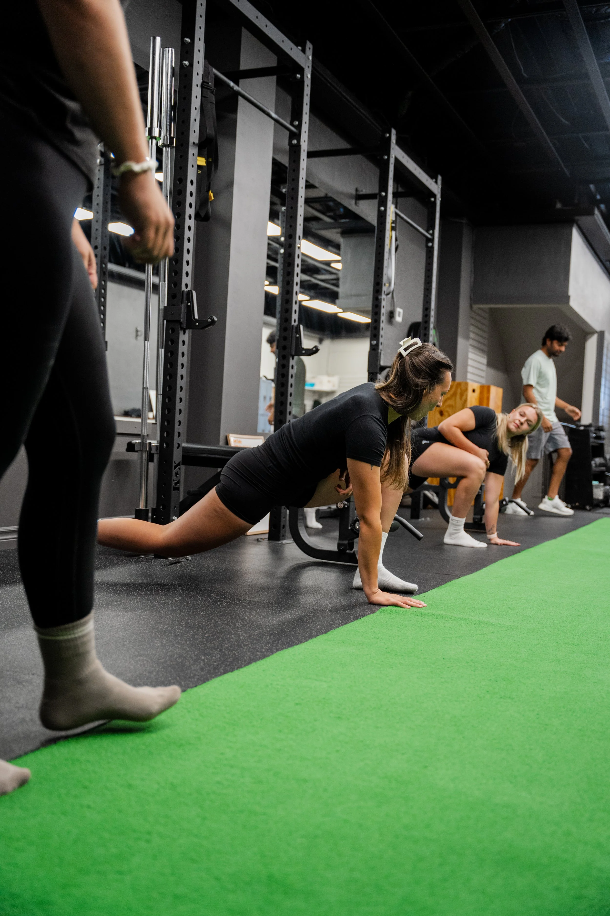 People exercising at a gym, with one woman performing a stretch on the floor and others in the background preparing for their workouts.
