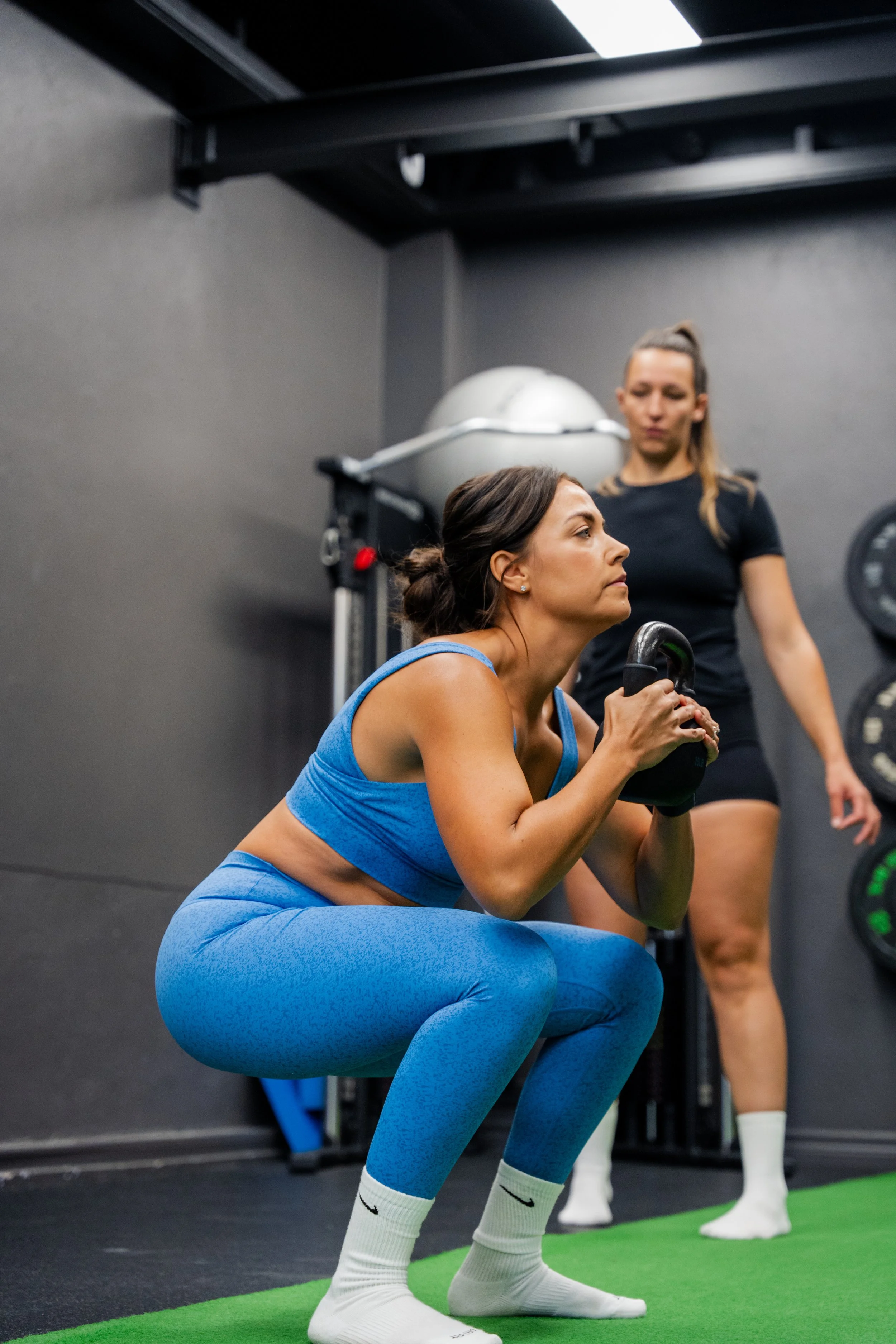 Woman in blue workout clothes in squat position lifting a kettlebell, with another woman standing behind her in a gym.