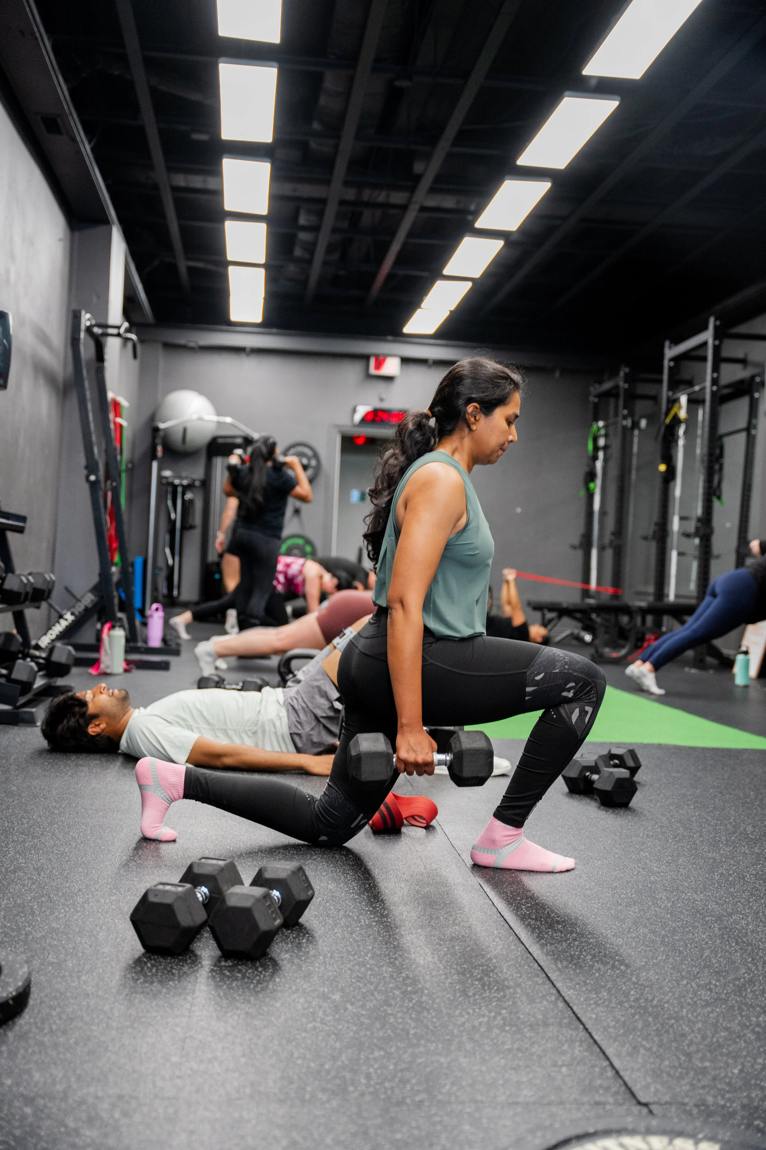 Woman performing lunges with dumbbells in a gym, with other people exercising in the background.