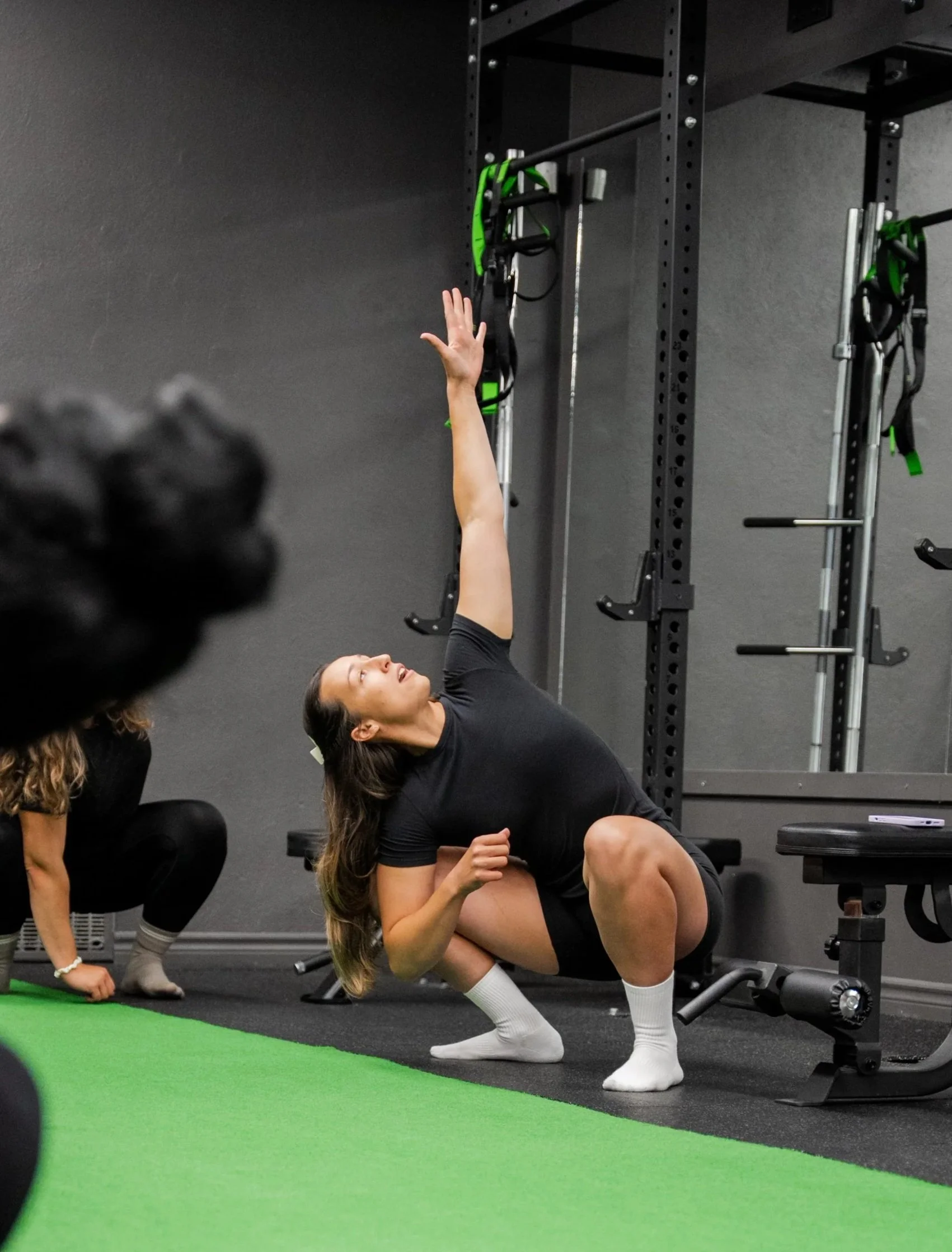 A woman doing a stretch or exercise on a black mat at the gym, with her right arm extended upward and her left arm resting on her bent left knee, next to workout equipment.