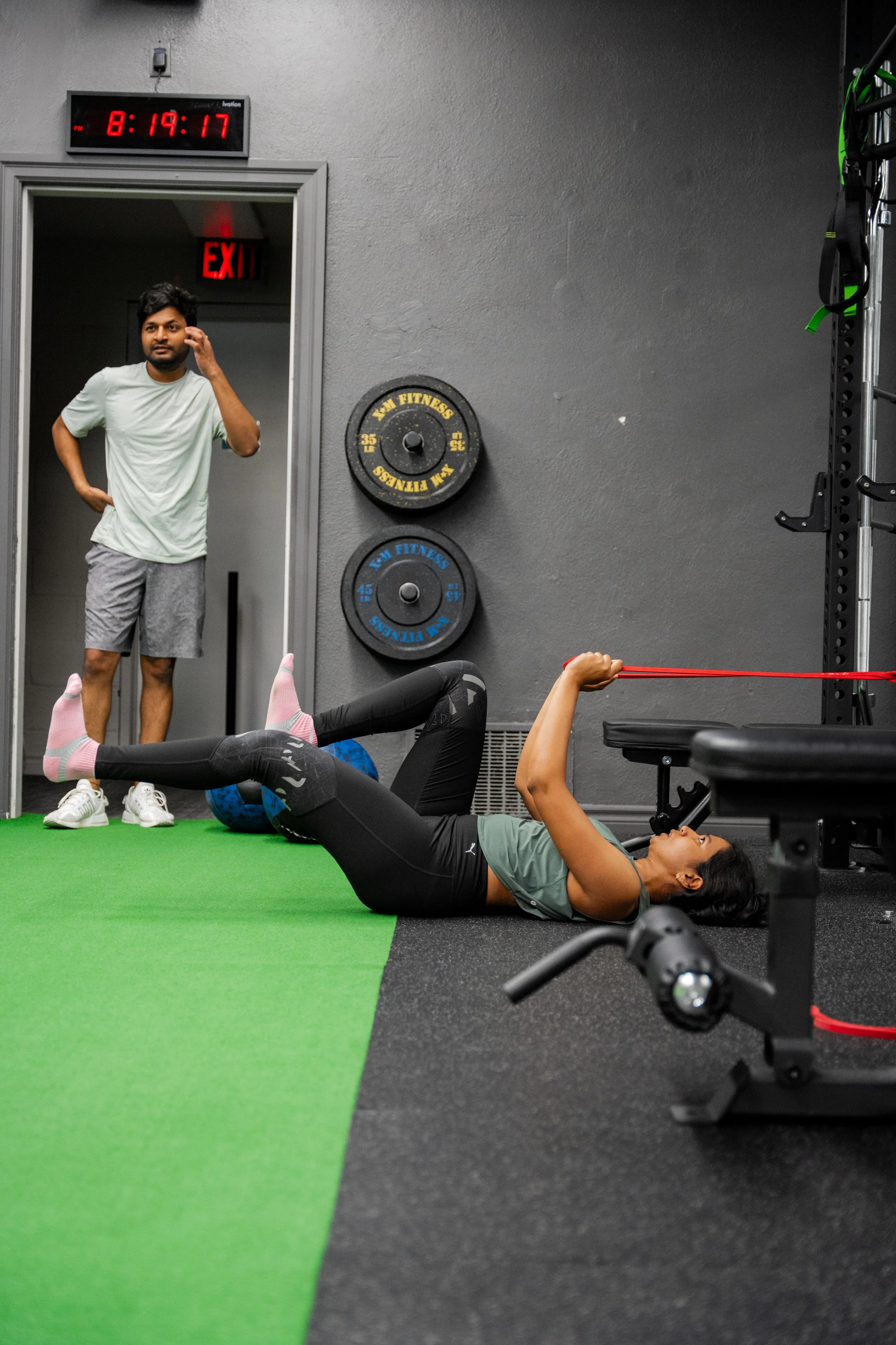 A woman exercises with resistance bands on her arms, lying on the gym floor, while a man watches nearby in a fitness gym with weights and equipment, and a digital clock shows the time as 8:19:17.