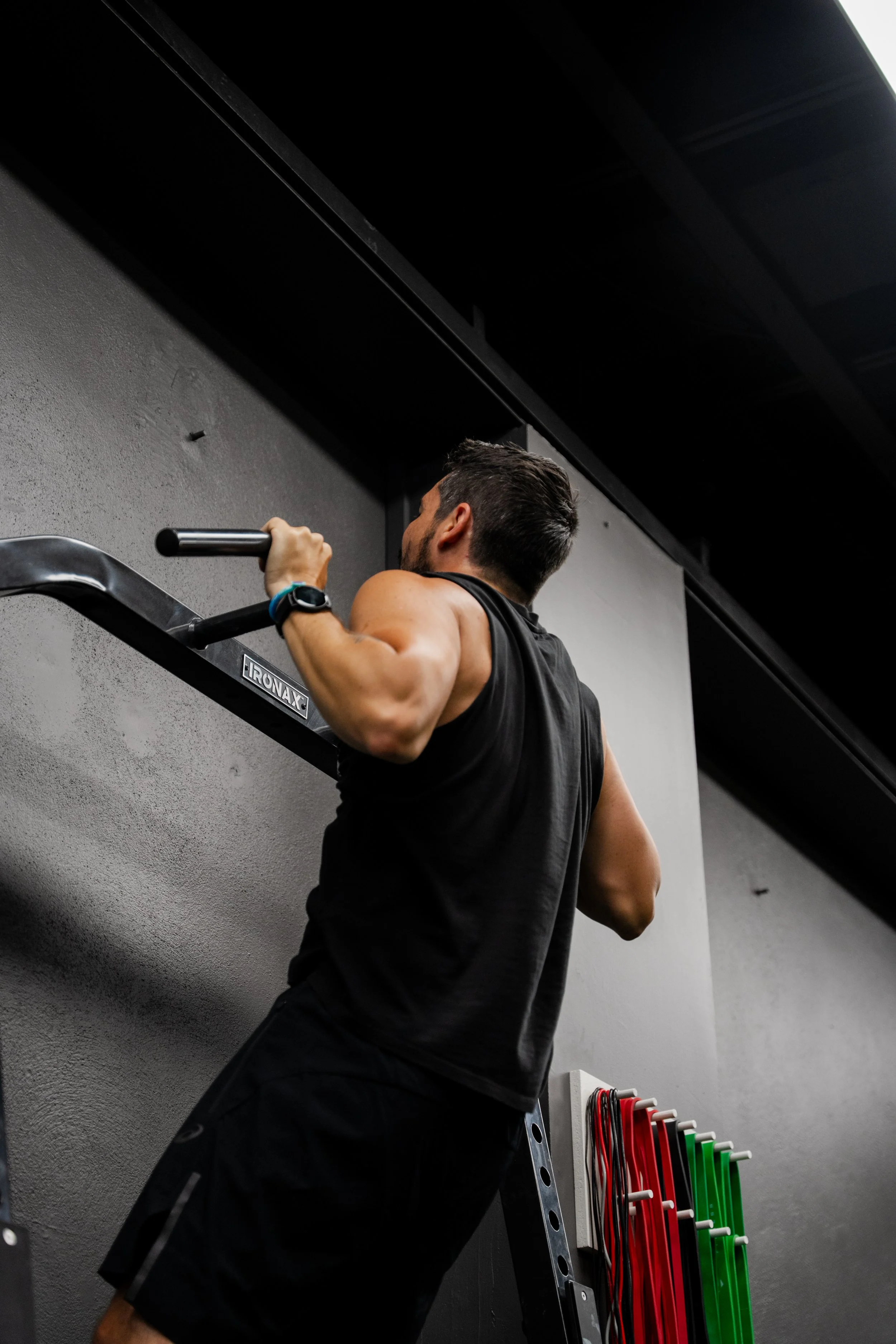 A man performs a pull-up exercise on a pull-up bar in a gym with gray walls.