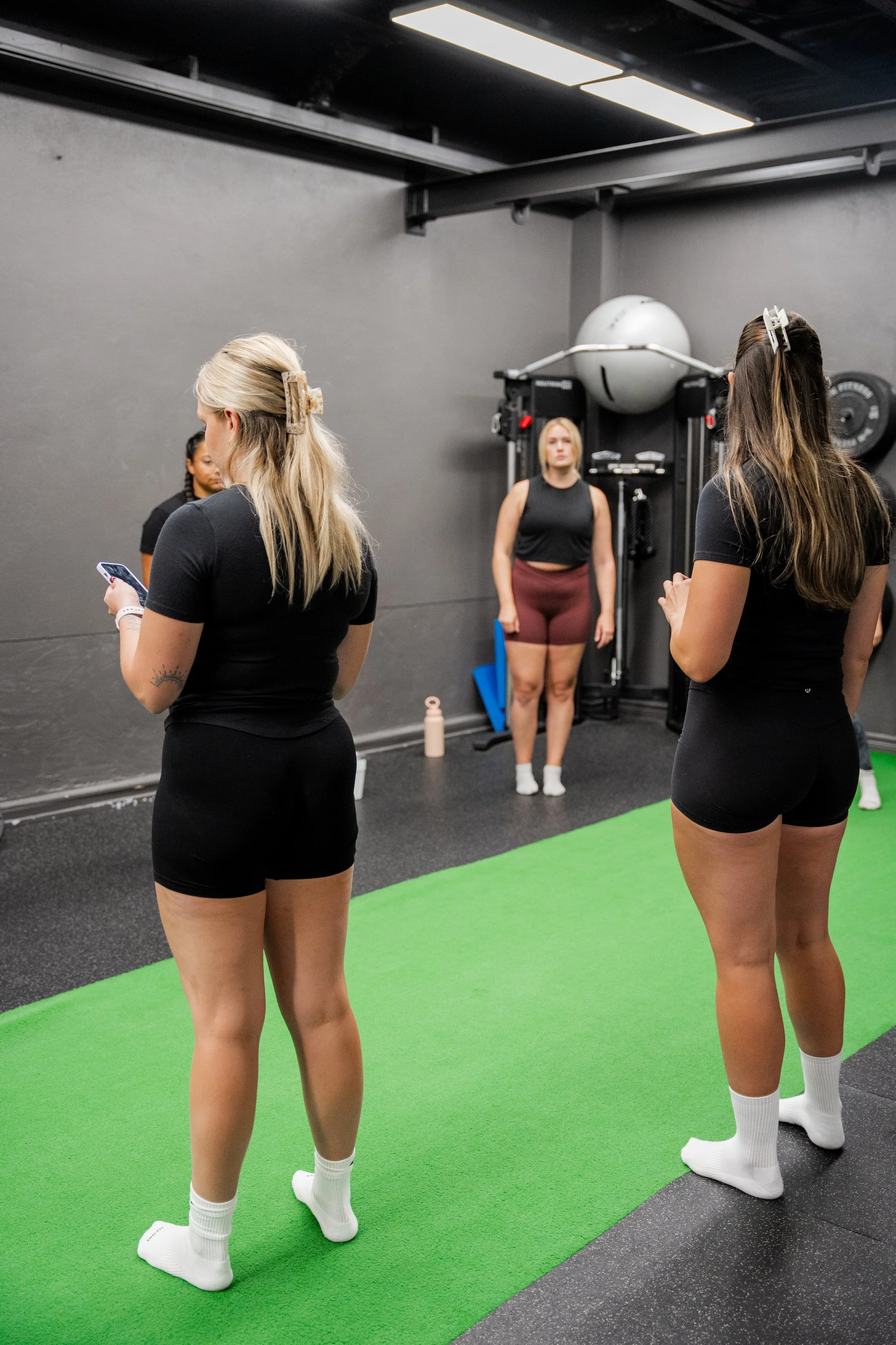 Four women with hair clips in their hair are standing on a green exercise mat in a gym, some are looking at their phones, while one woman is facing the camera. The gym has black walls and fitness equipment in the background.