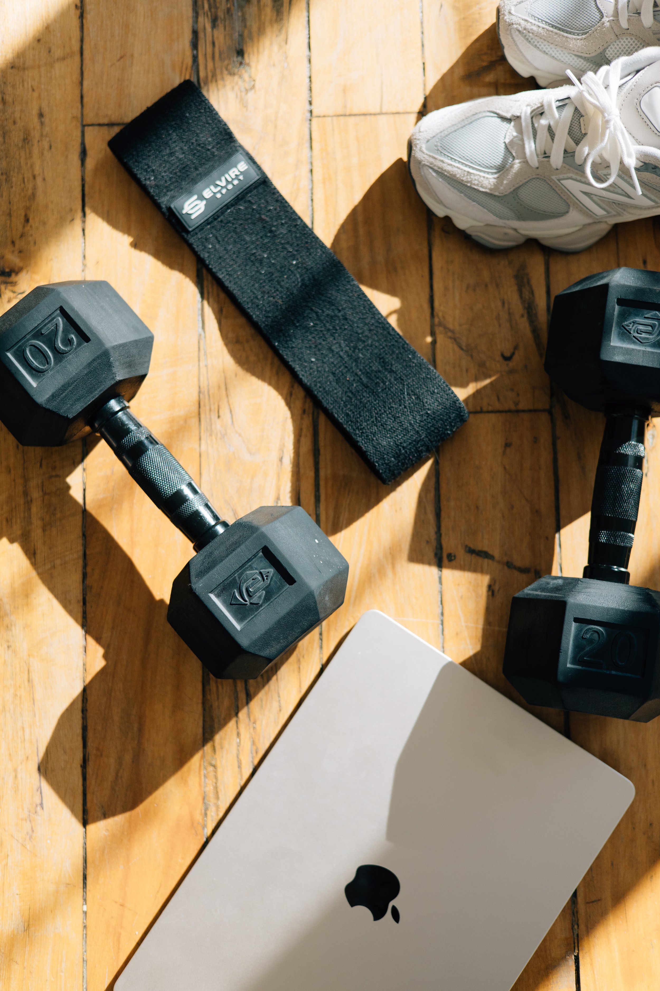 Dumbbells, a wrist strap, a white athletic shoe, a closed laptop, and a fitness tracker are laid out on a wooden floor.