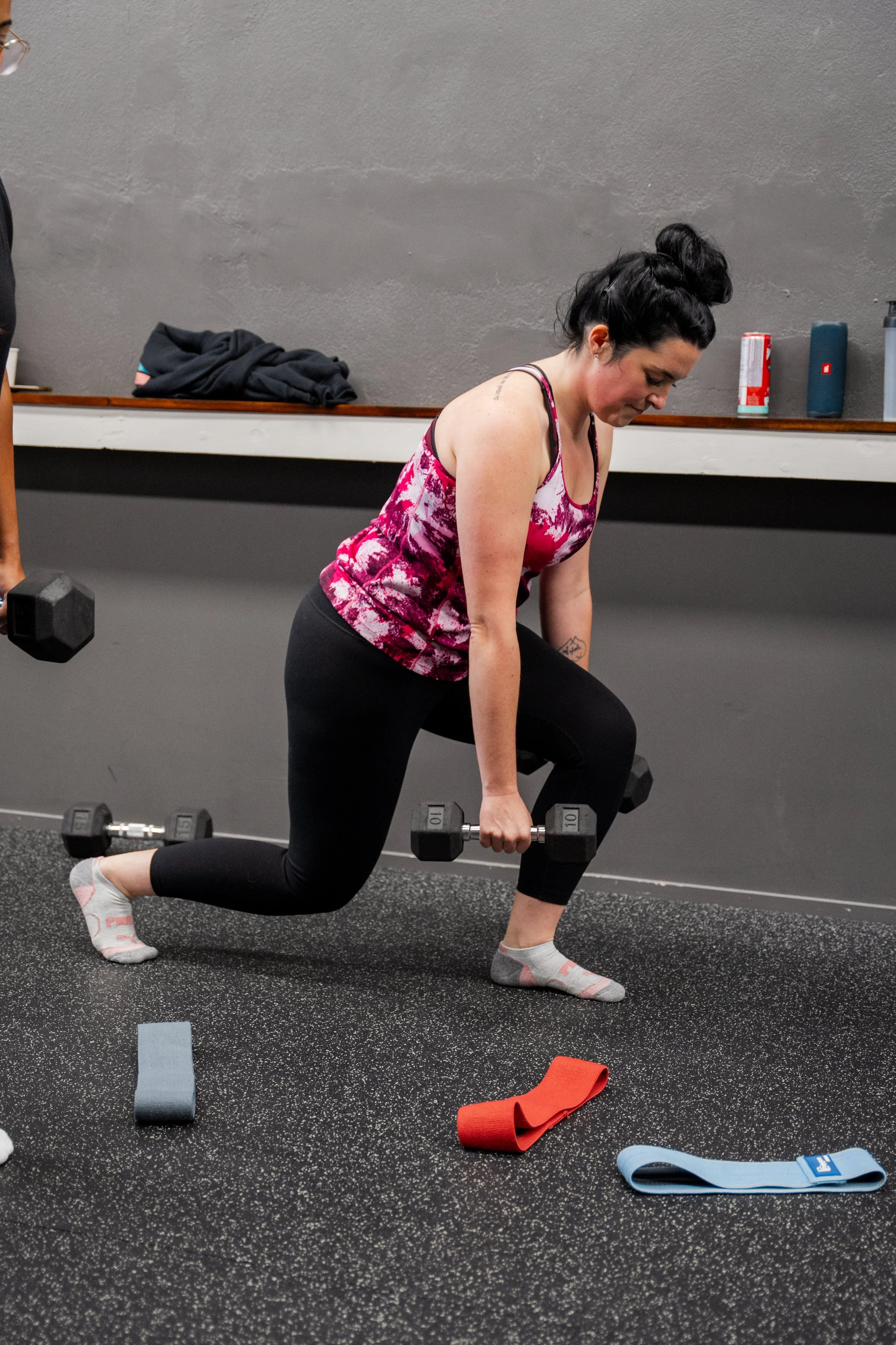 A woman in workout clothes lifting weights while one knee is on the ground in a gym.