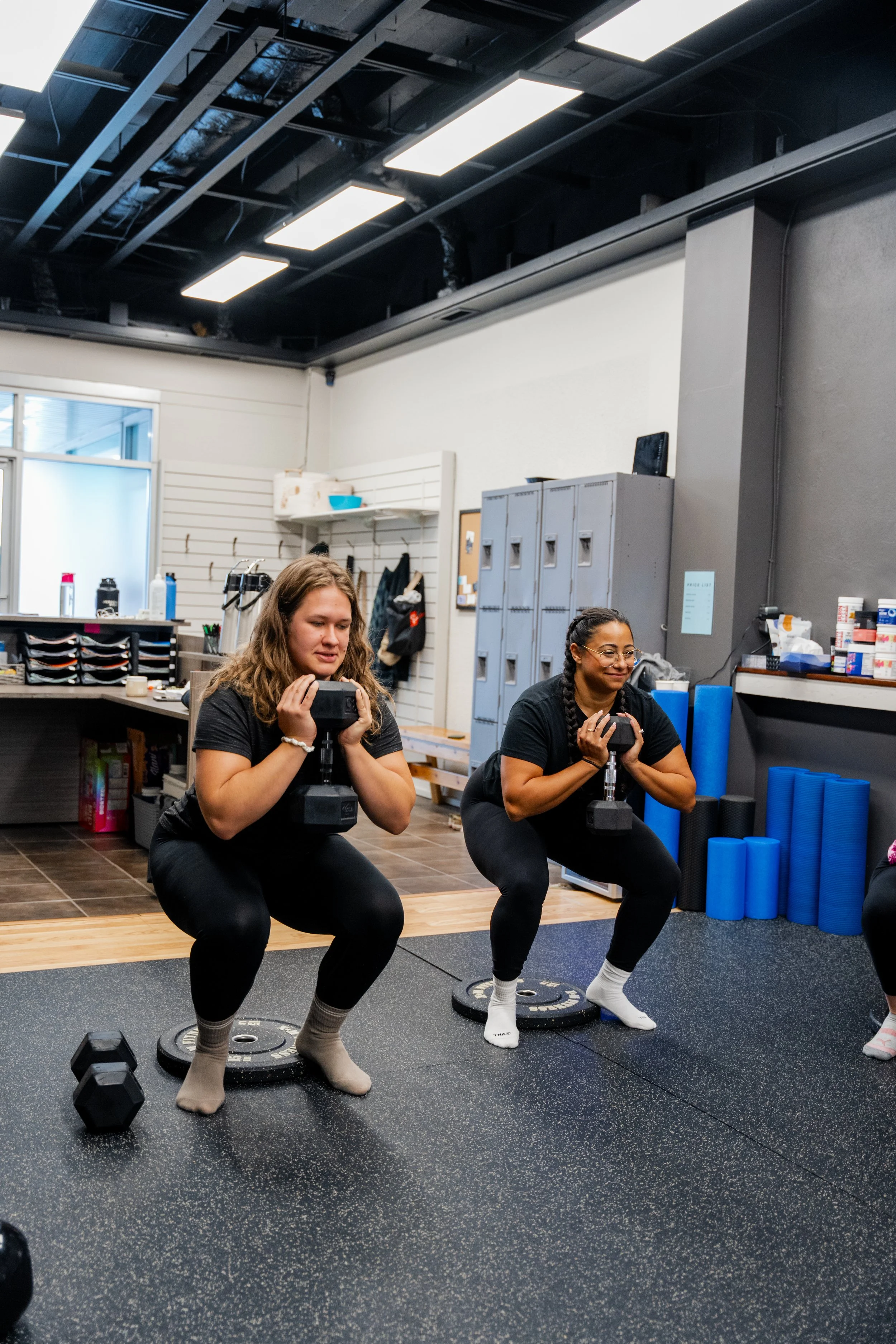 Two women with long hair, wearing black t-shirts and black leggings, are exercising with dumbbells in a gym. They are squatting on weight plates placed on the floor, and both are smiling and focused. The gym has lockers, foam rollers, and other exerc