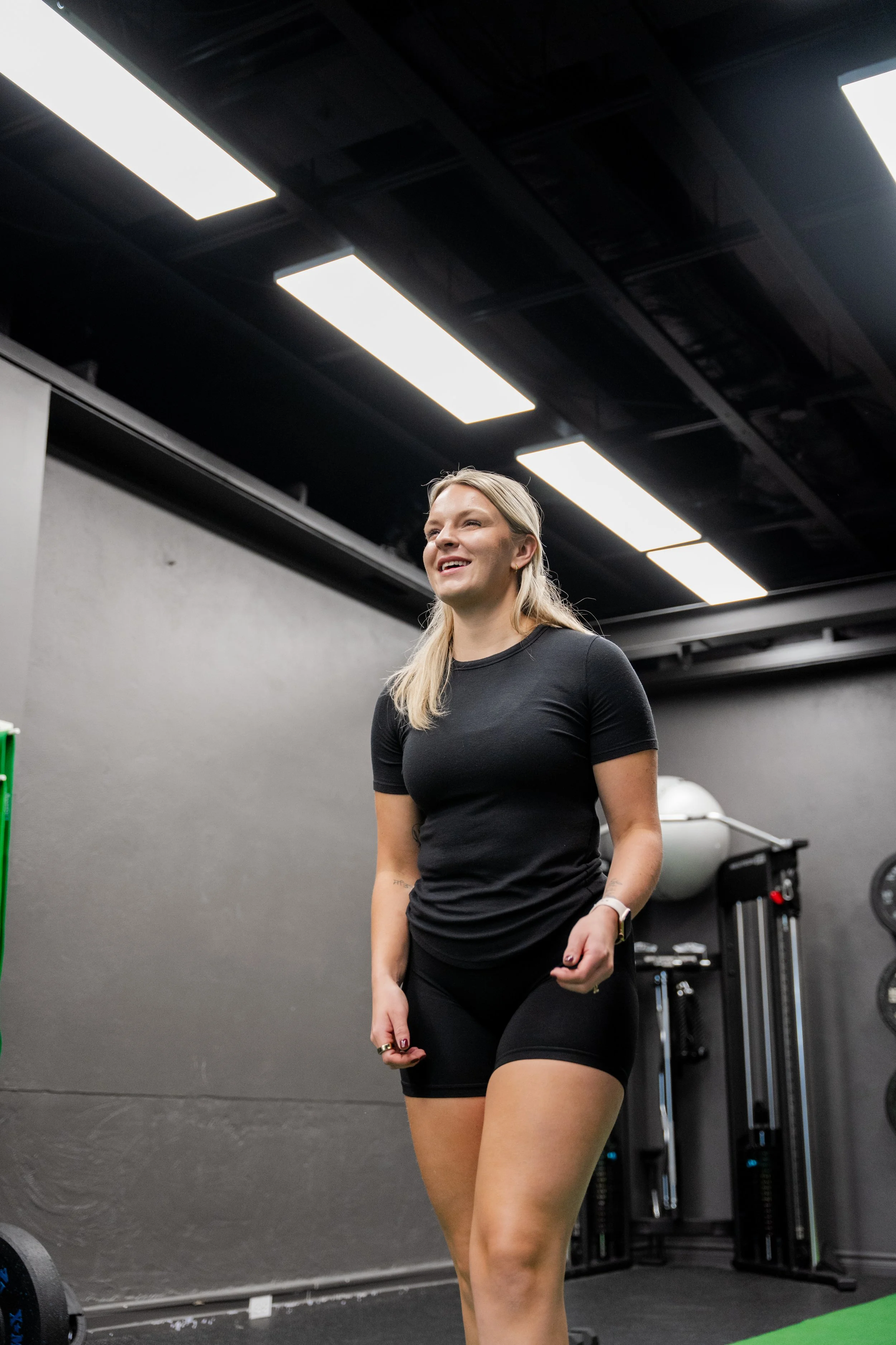 A woman in workout clothes standing in a gym, smiling with her fists slightly clenched.