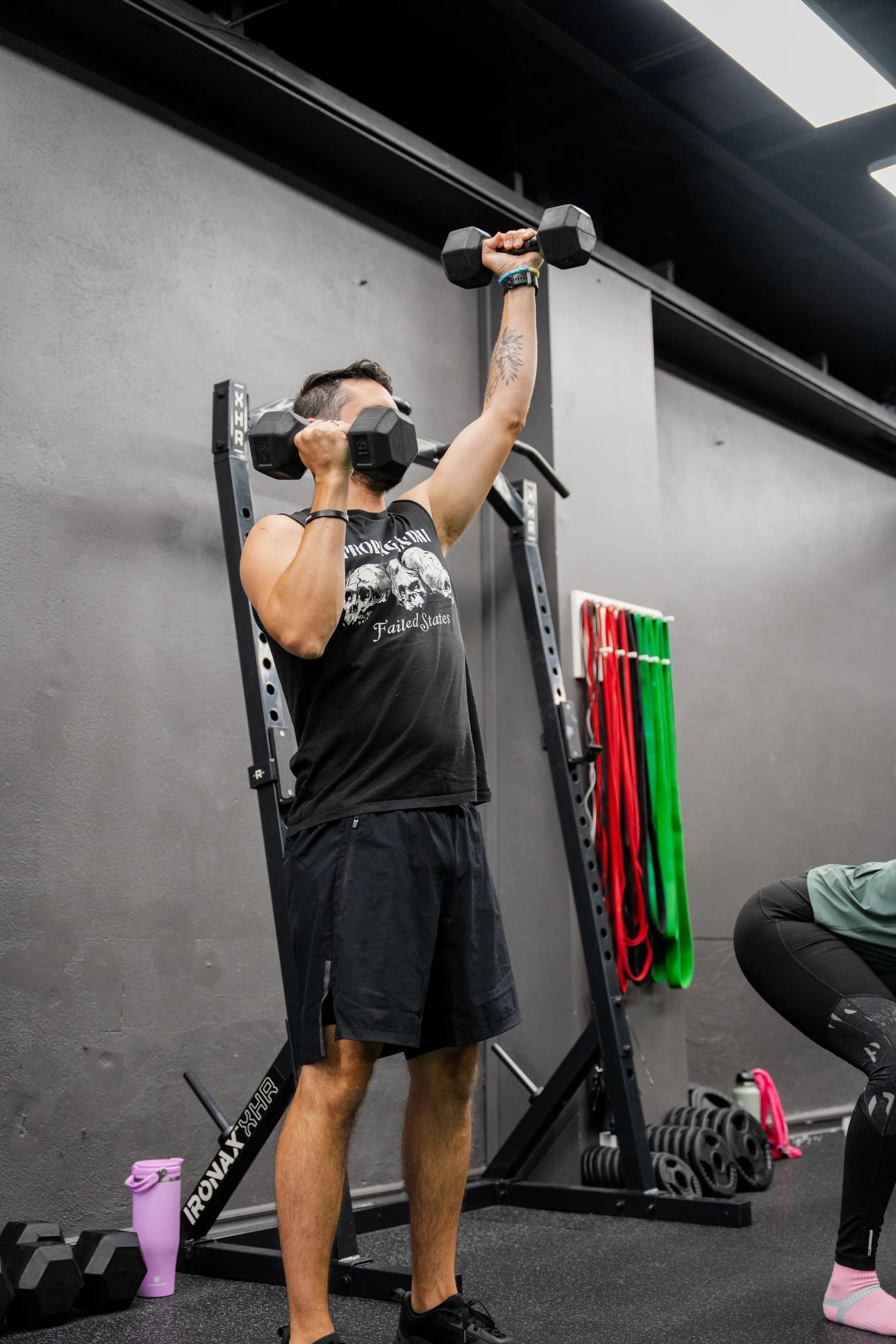 A man in a black tank top and shorts exercises with a dumbbell in a gym, raising it above his head.