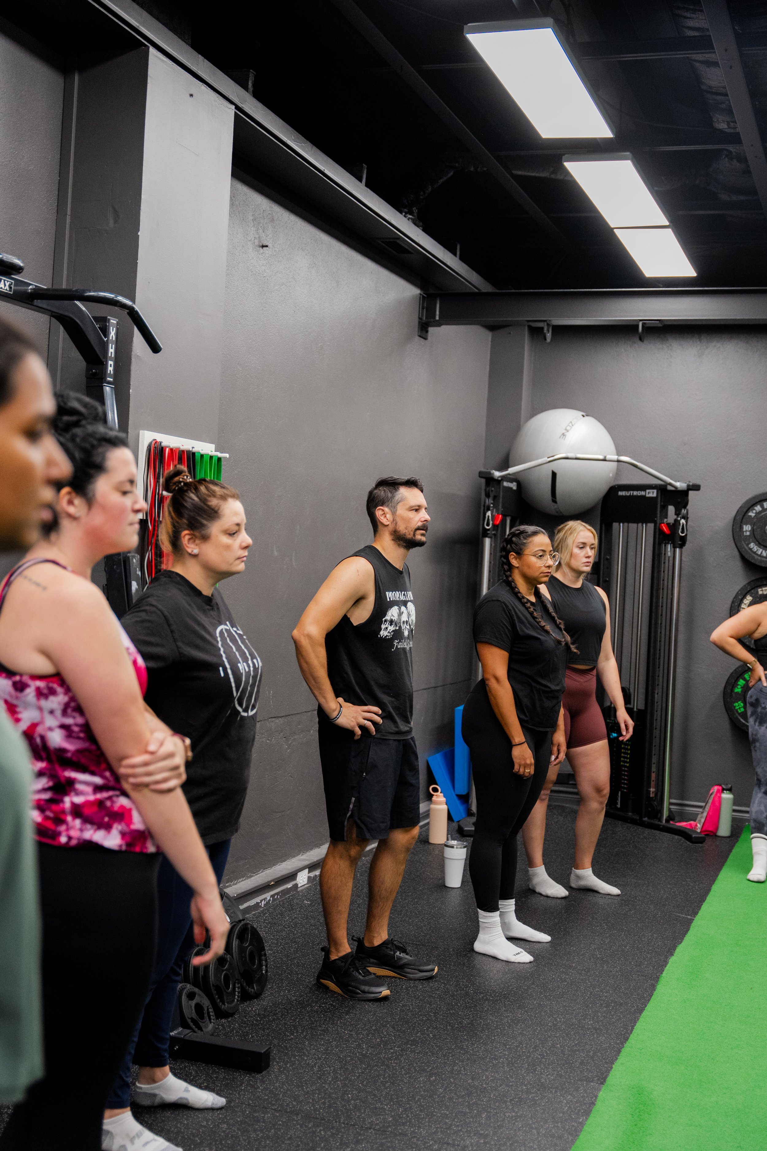 Group of people standing in a gym, paying attention to someone outside the frame, with workout equipment like resistance bands and a stability ball visible.