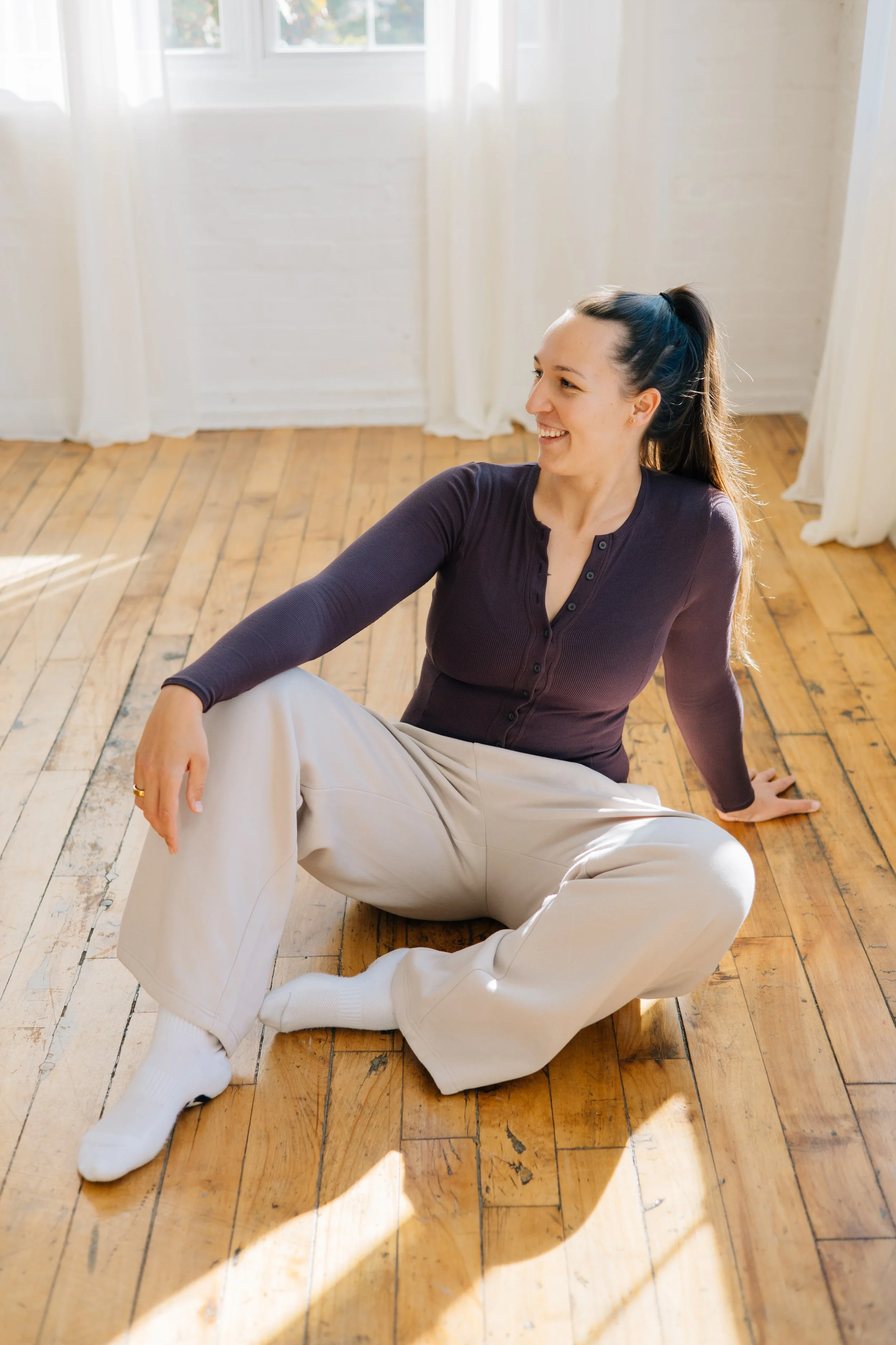 A woman sitting on a wooden floor, smiling and looking to her right, with sunlight coming through white curtains in the background.