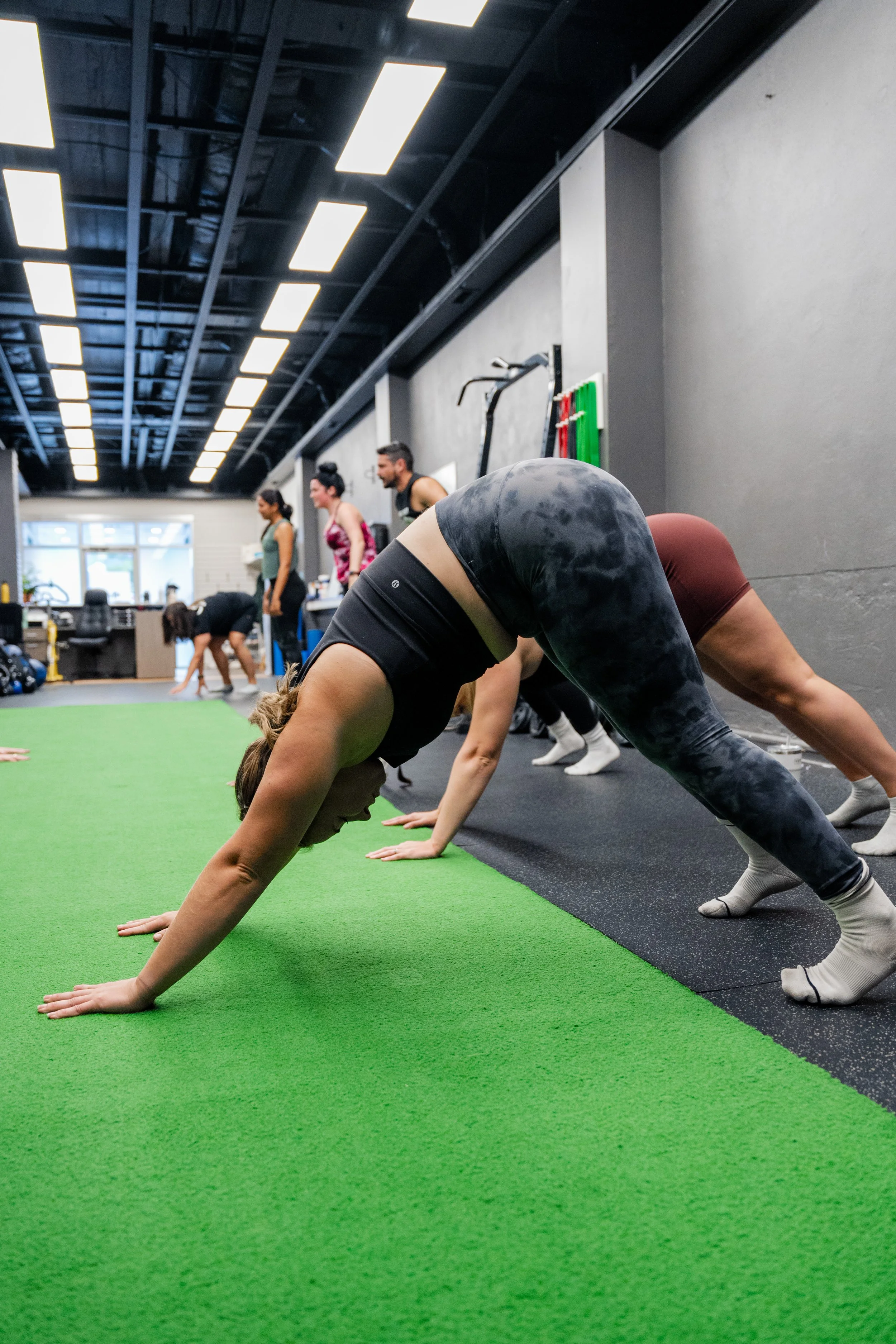People in a gym practicing yoga on the green mat, with a woman in the foreground in a downward dog pose.