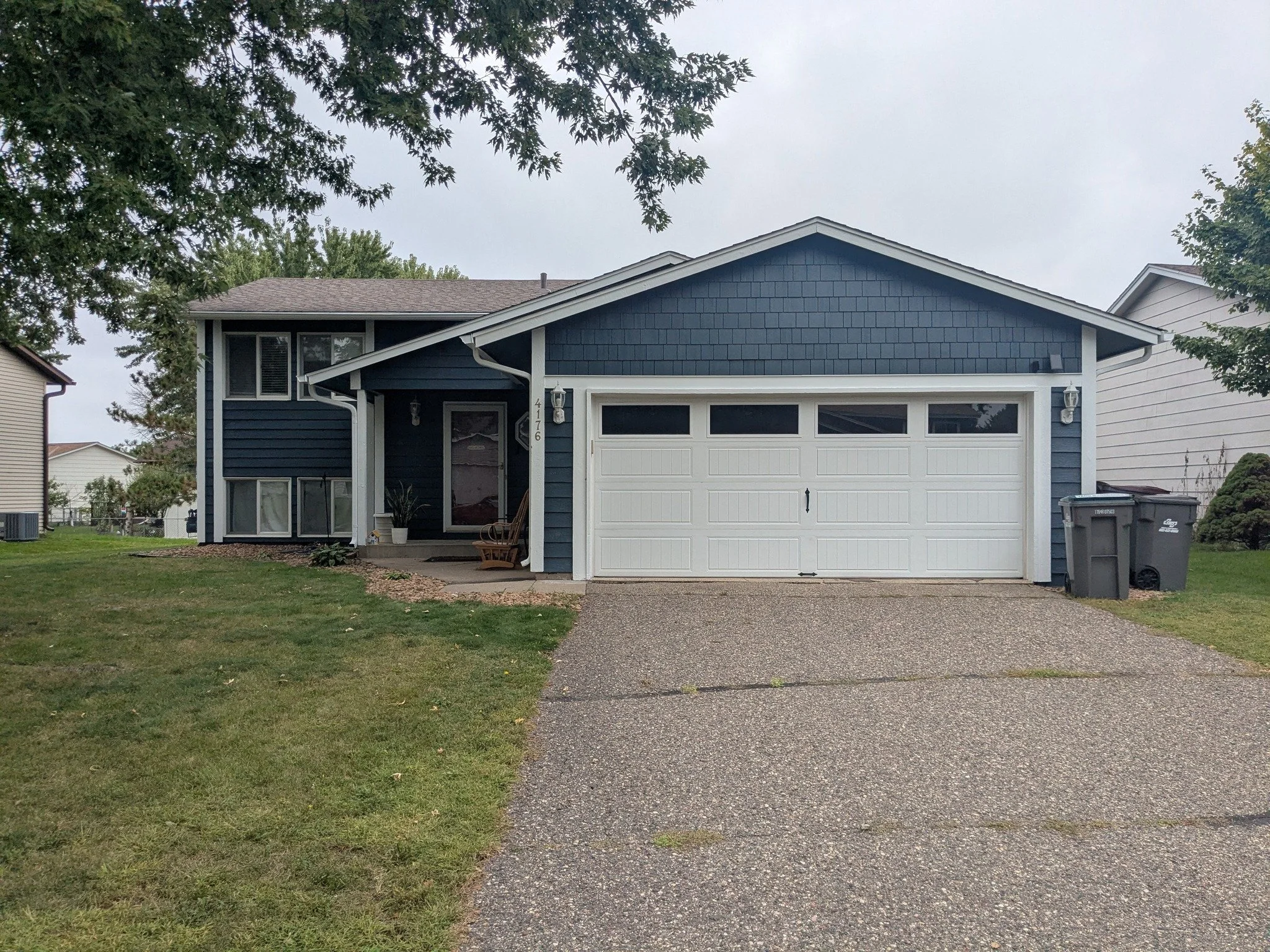 A two-story blue house with a white garage door and a small front porch. The house has a dark roof and is surrounded by a grassy lawn, with trees on the sides. There are two trash bins next to the garage and a rocking chair near the front door.