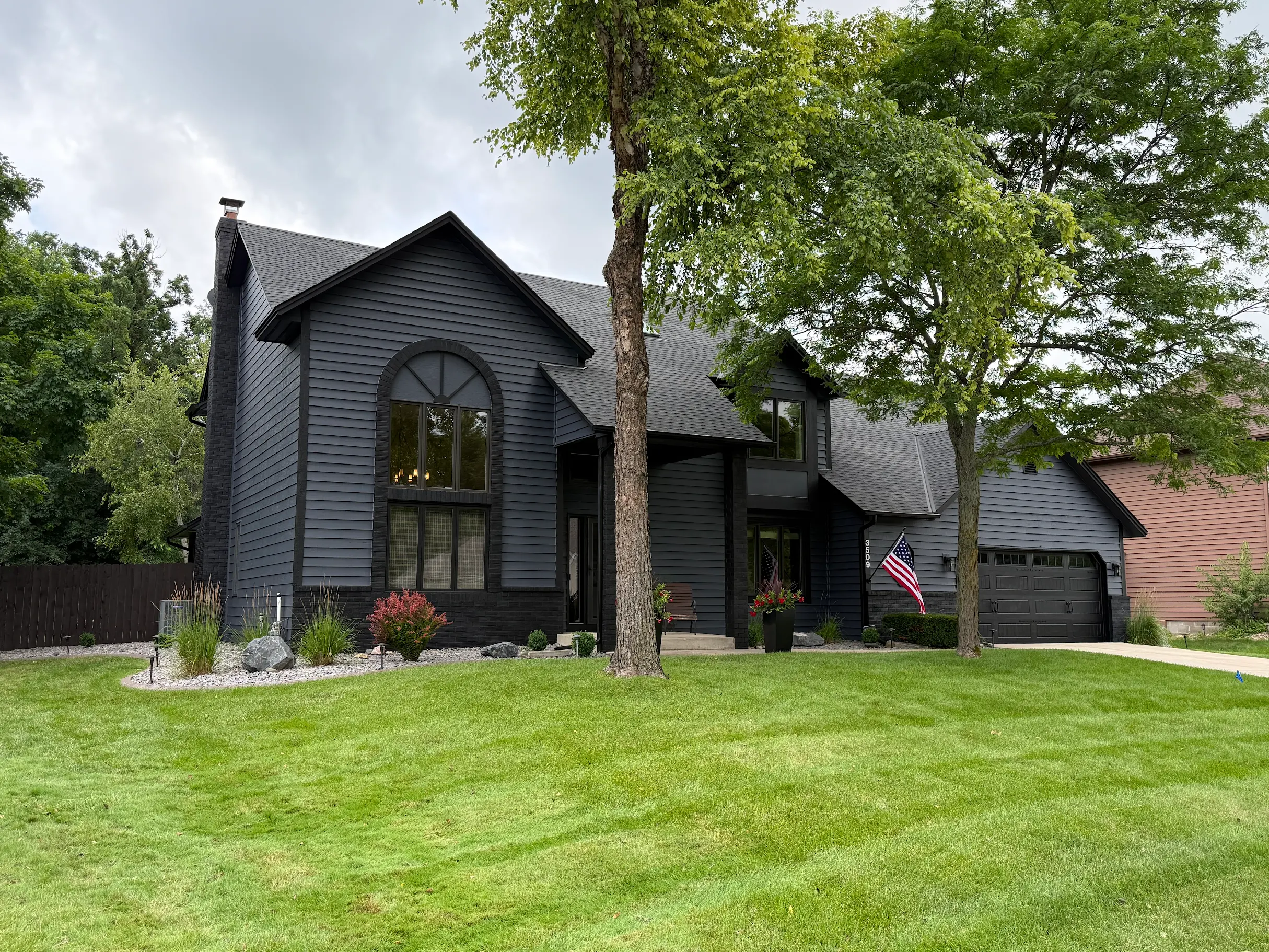 A dark gray two-story house with black trim, large front window, attached garage, and front porch with American flag, surrounded by green lawn, trees, and landscaped garden beds.
