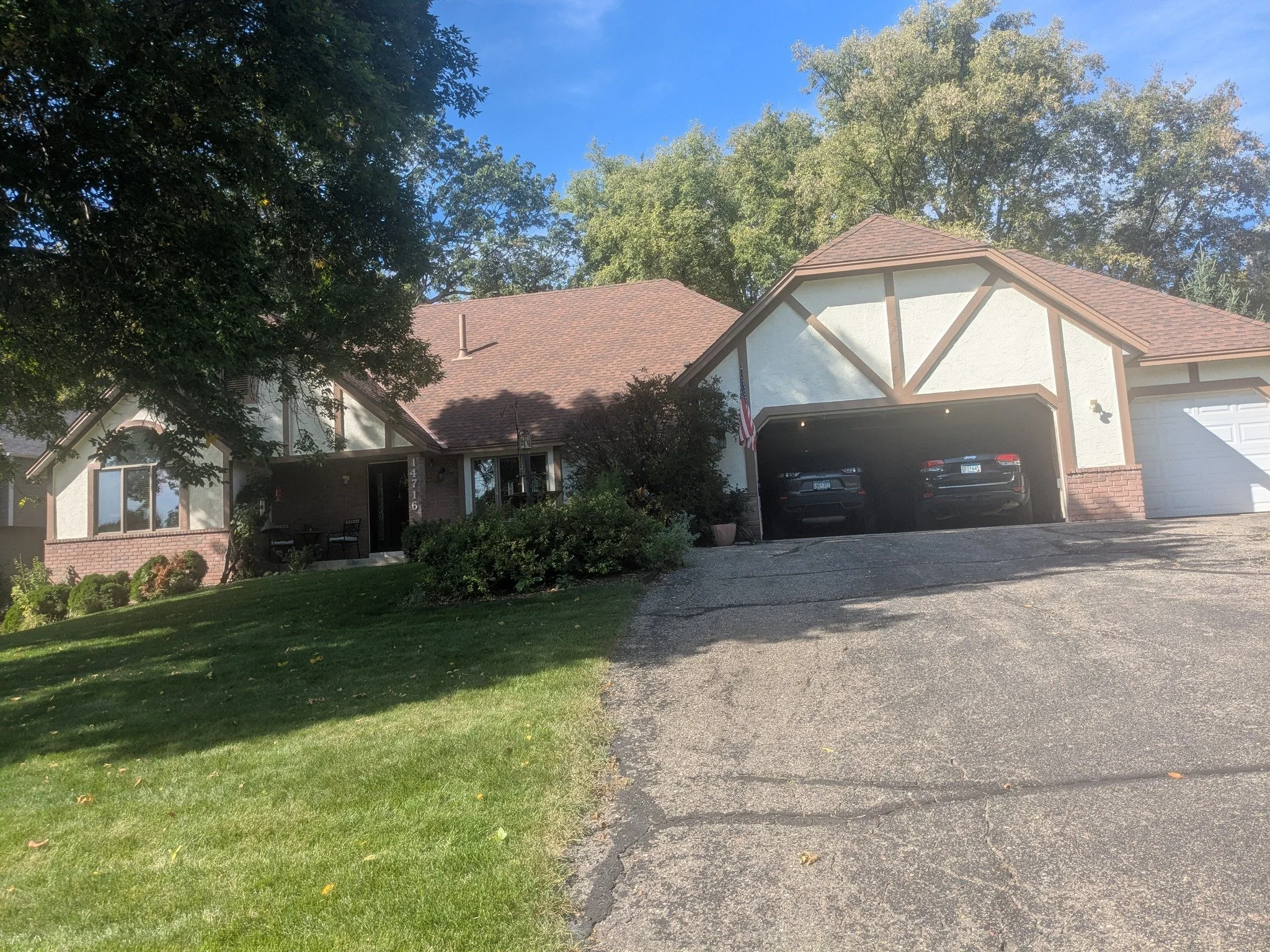 A two-story house with a sloped roof, a two-car garage, and a front yard with green grass and bushes. The house has white walls with brown wooden trim and an American flag hanging near the garage. Trees are in the background and the sky is clear and blue.