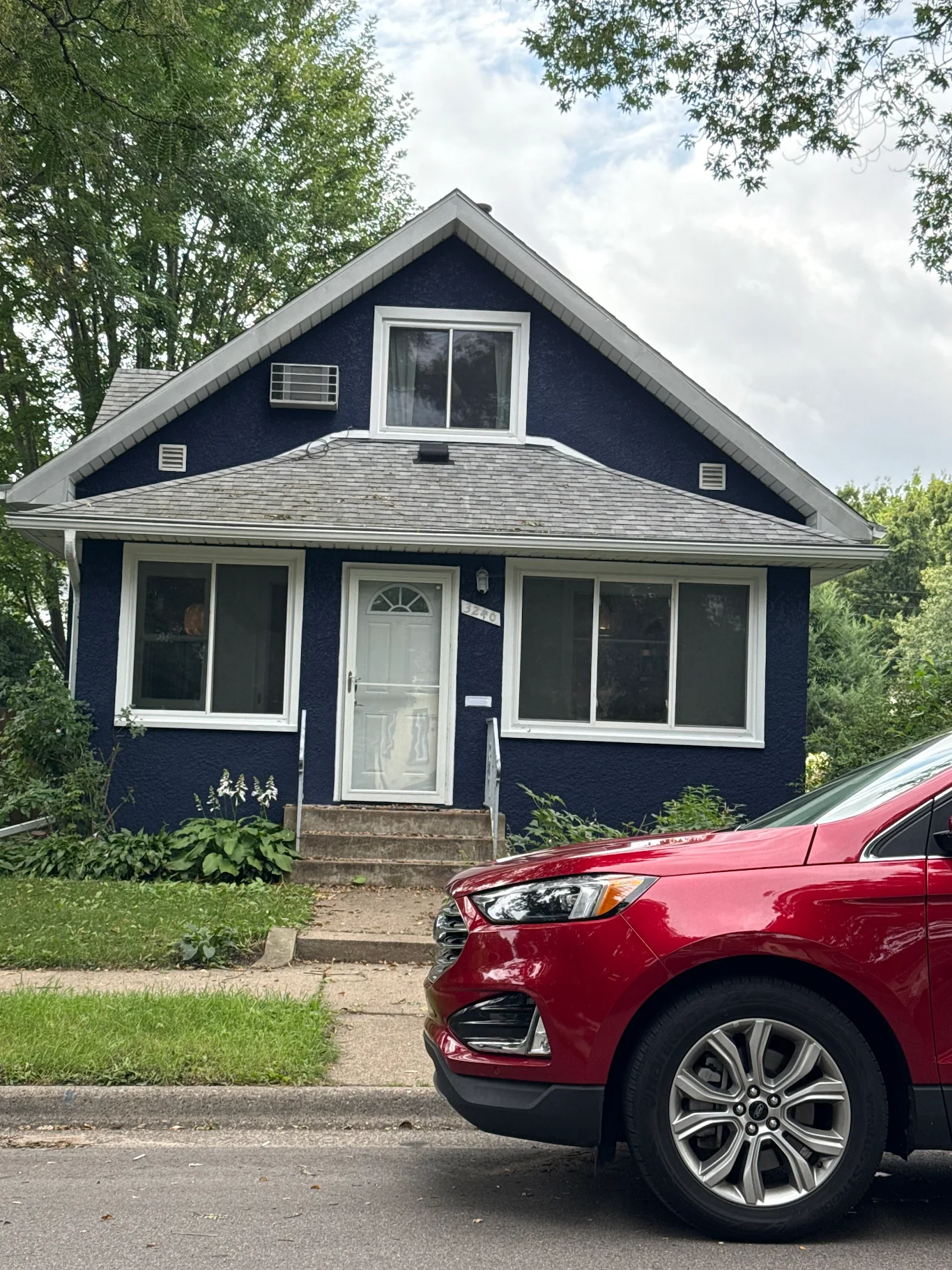 A blue house with white trim and a gabled roof, with a small front porch and steps. There is a red car parked in front of the house on the street. The house has large windows on either side of the front door and a window on the second floor. There are trees and greenery around the house.