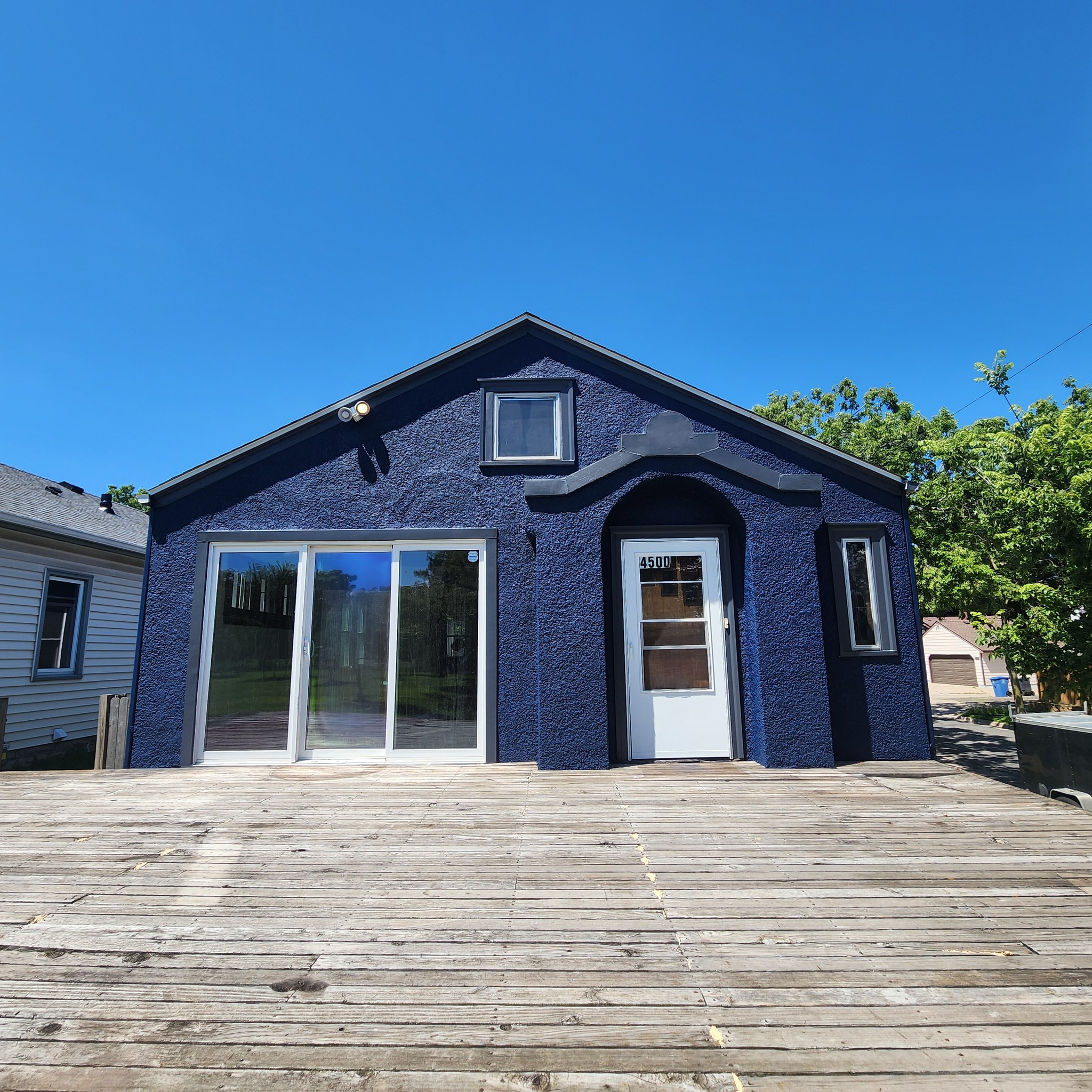 A small, blue house with a textured exterior, large sliding glass door, a white front door with the number 4500, and a small window above it, set against a bright blue sky with some green trees on the side.