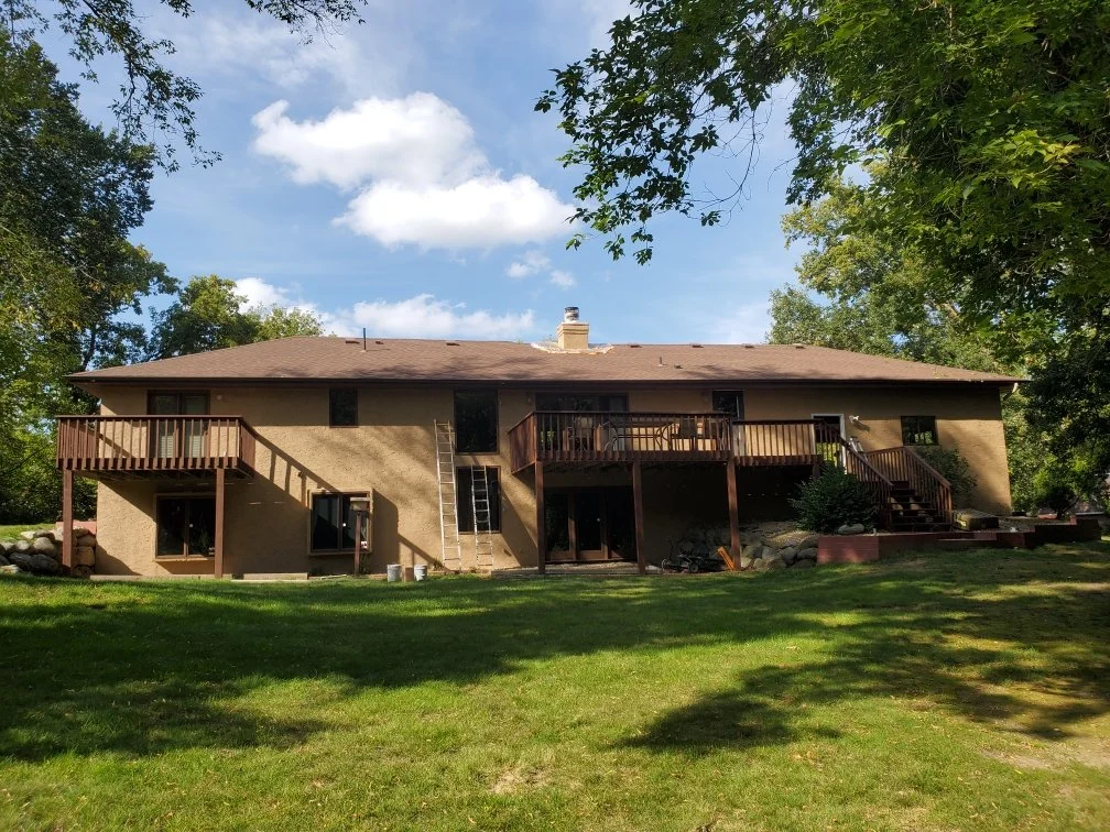 Back view of a two-story house with a brown roof and exterior walls, featuring two wooden decks on the upper level, surrounded by green trees and grass, under a blue sky with some clouds.