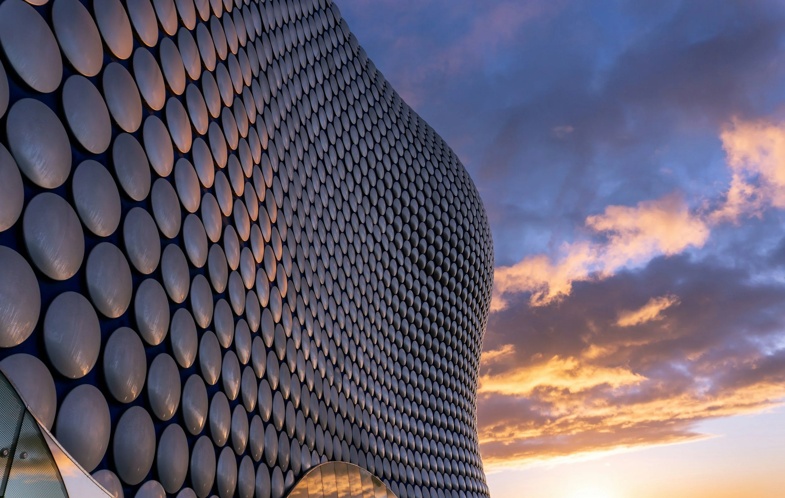 A modern building with a curved facade covered in circular, metallic panels during a sunset with partly cloudy sky.