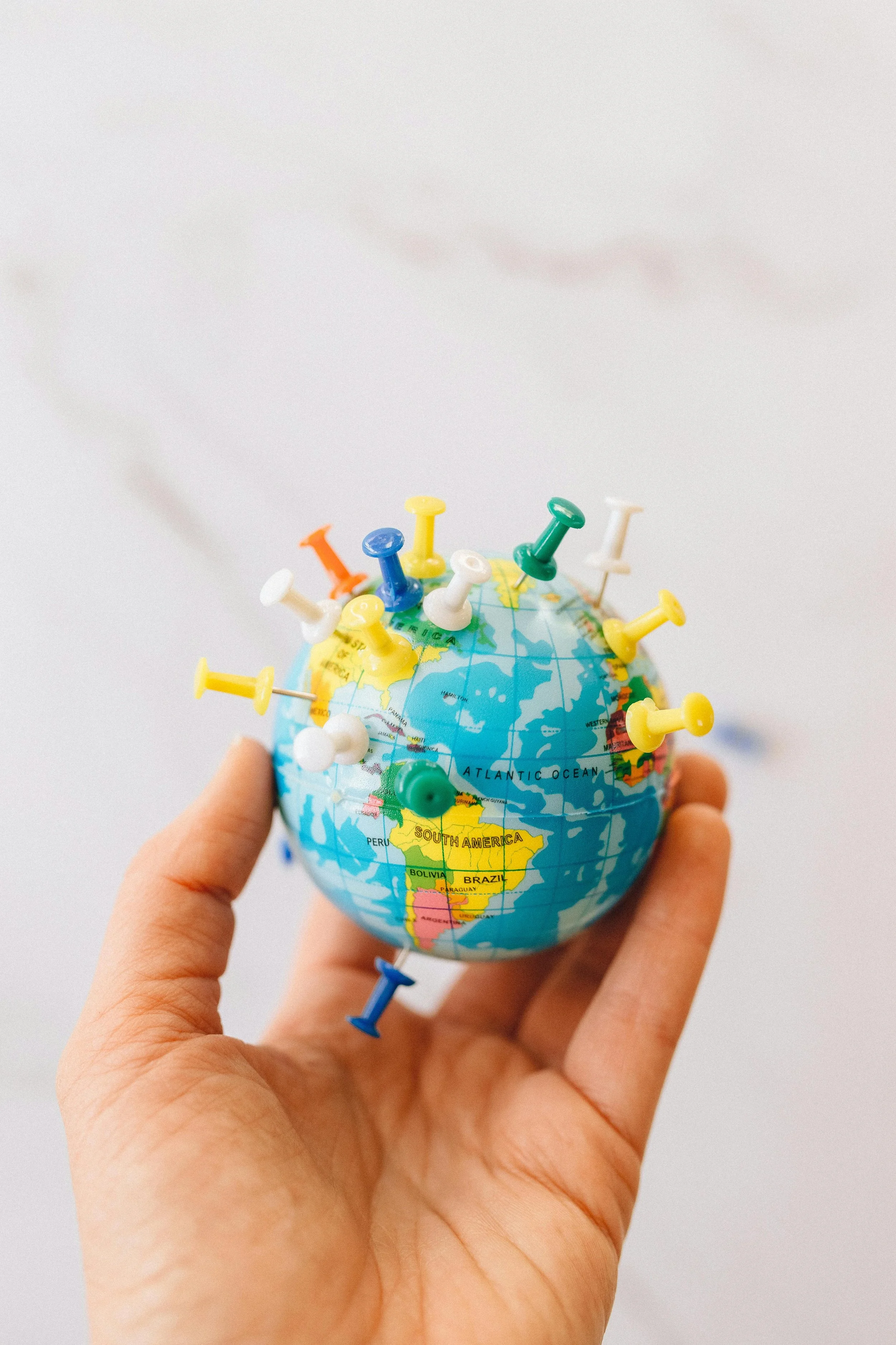 A small globe with colorful pushpins stuck in various locations, held in a person's hand against a plain background.