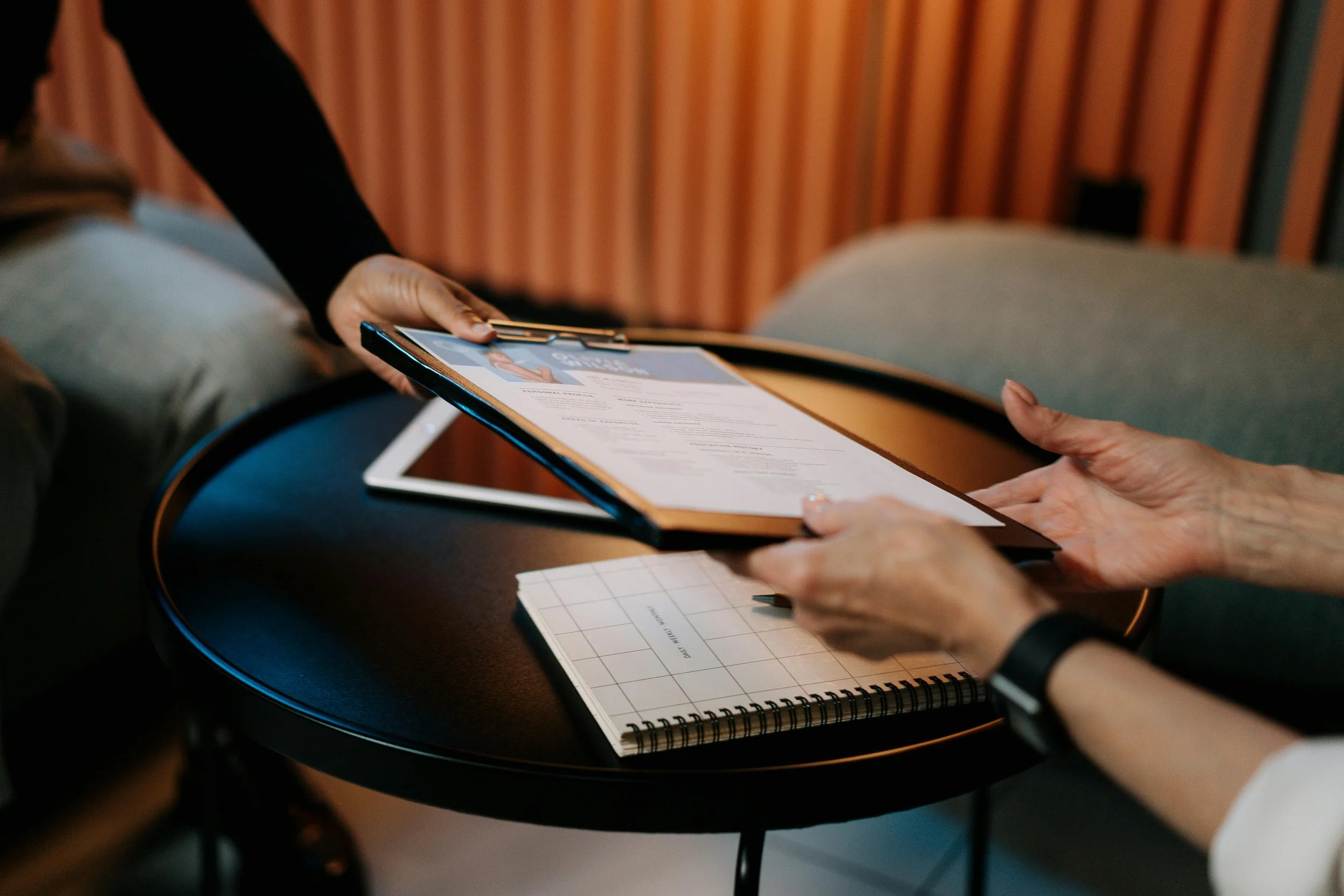 Two people exchanging a document or certificate at a small round table, with a notebook and a tablet present.