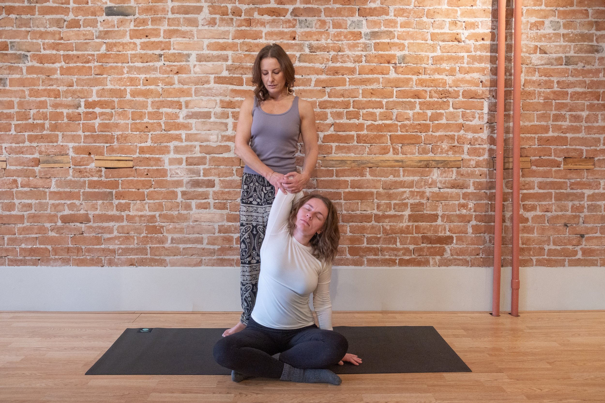 Two women practicing partner yoga or stretching in a studio with a brick wall background. One woman is seated on a yoga mat with her eyes closed, and the other is standing, holding her arm above her head.
