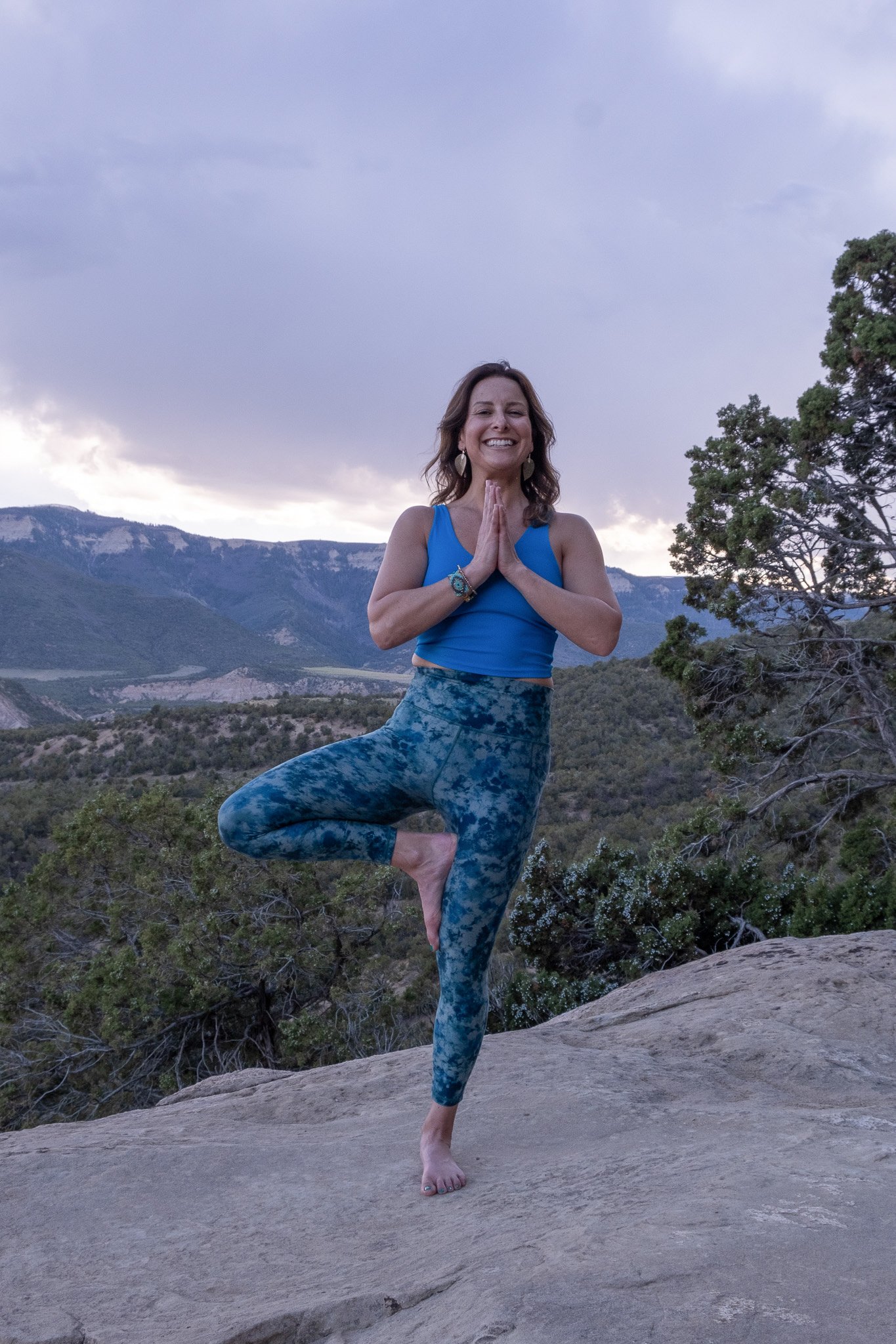 A woman practicing yoga outdoors on a rocky surface during sunset, standing in the tree pose with hands in prayer position, mountains and trees in the background.