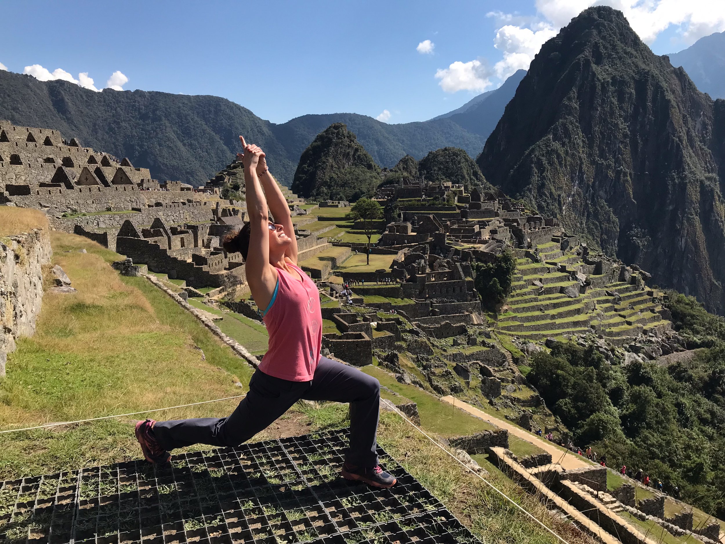 A woman doing yoga on a platform at Machu Picchu, Peru, with ancient stone ruins and mountain scenery in the background.