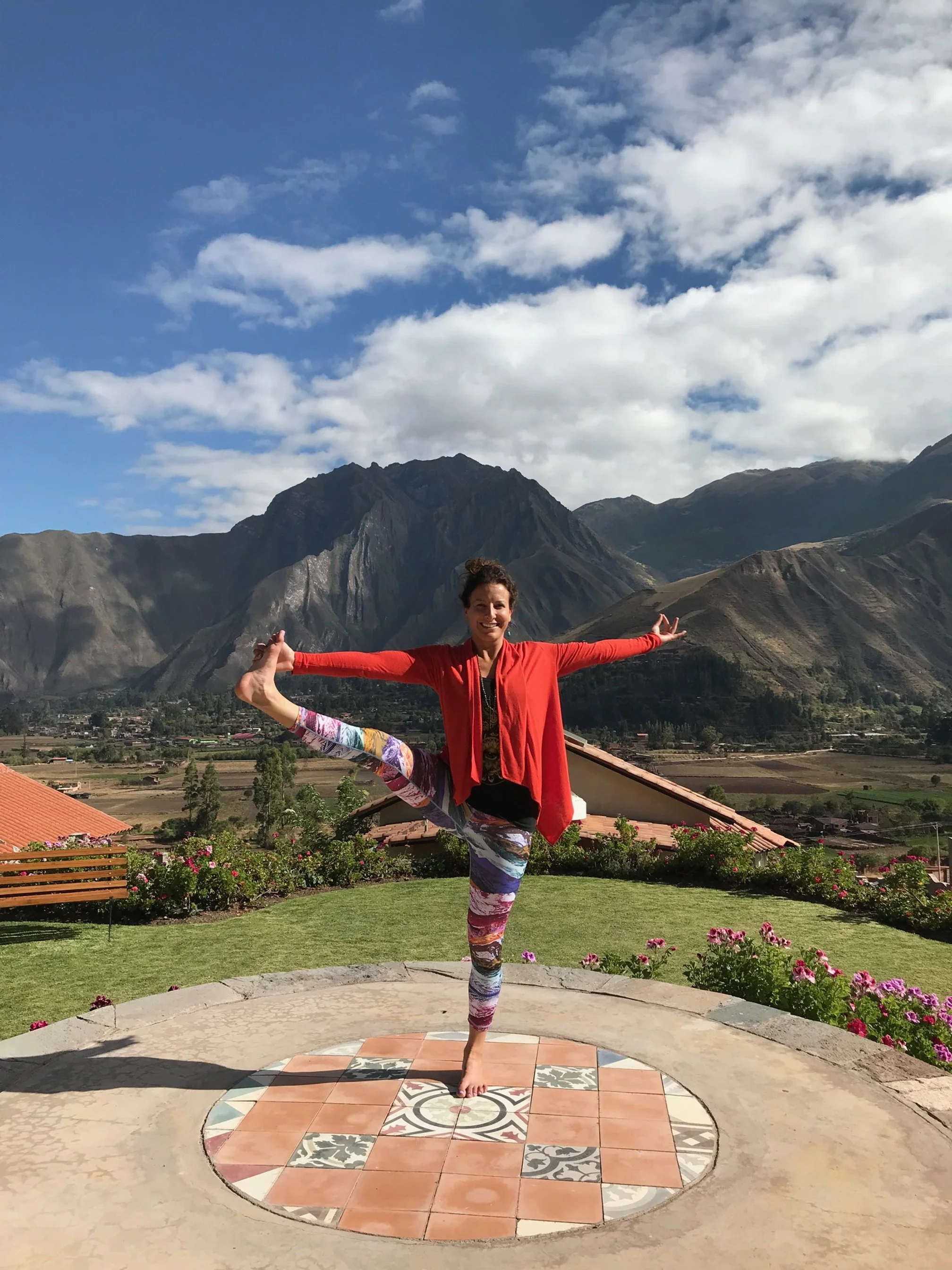 A woman practicing yoga outdoors on a tiled circular platform, with mountains and a partly cloudy sky in the background.