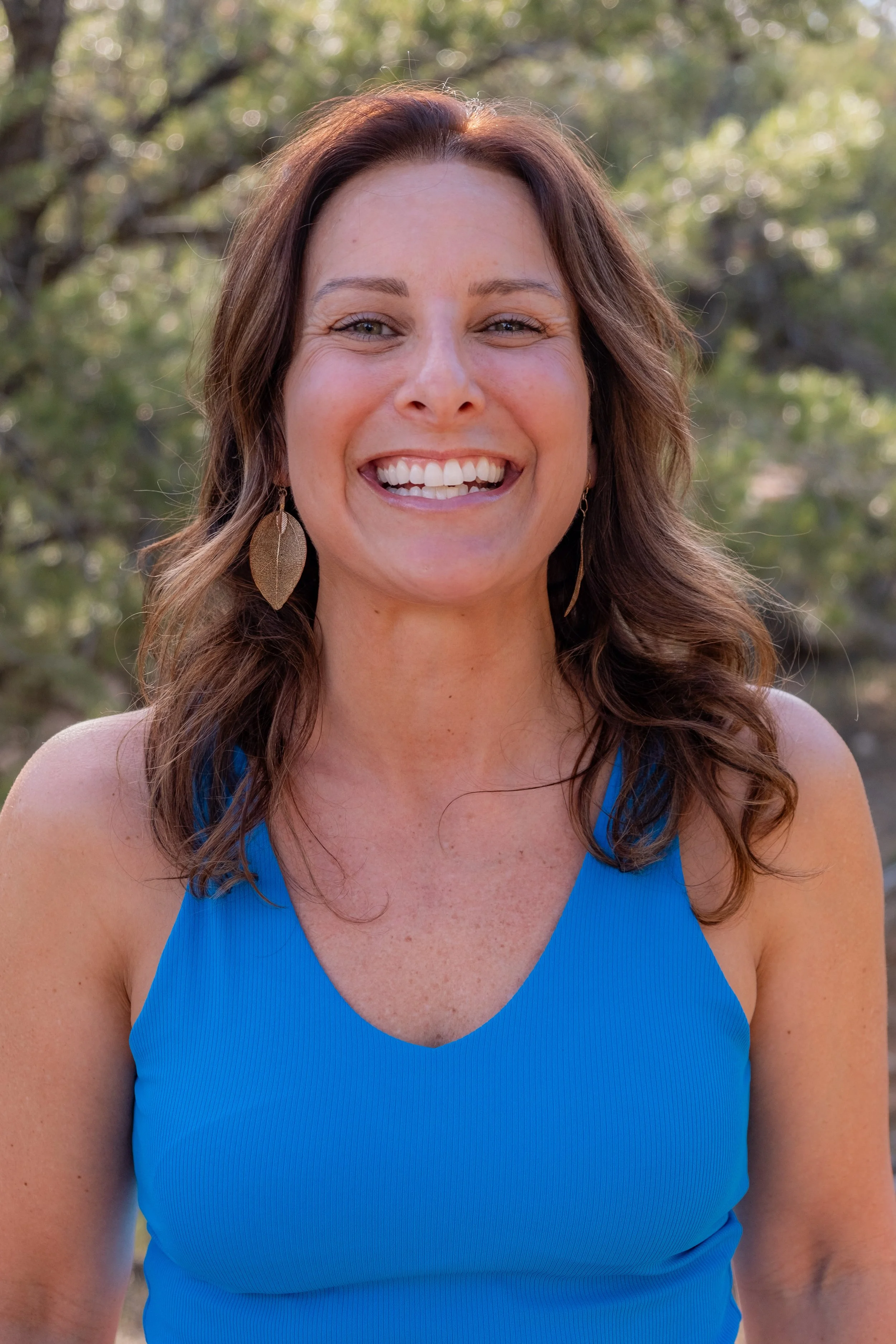 Smiling woman with brown hair wearing a blue sleeveless top and gold leaf earrings outdoors with trees in the background.