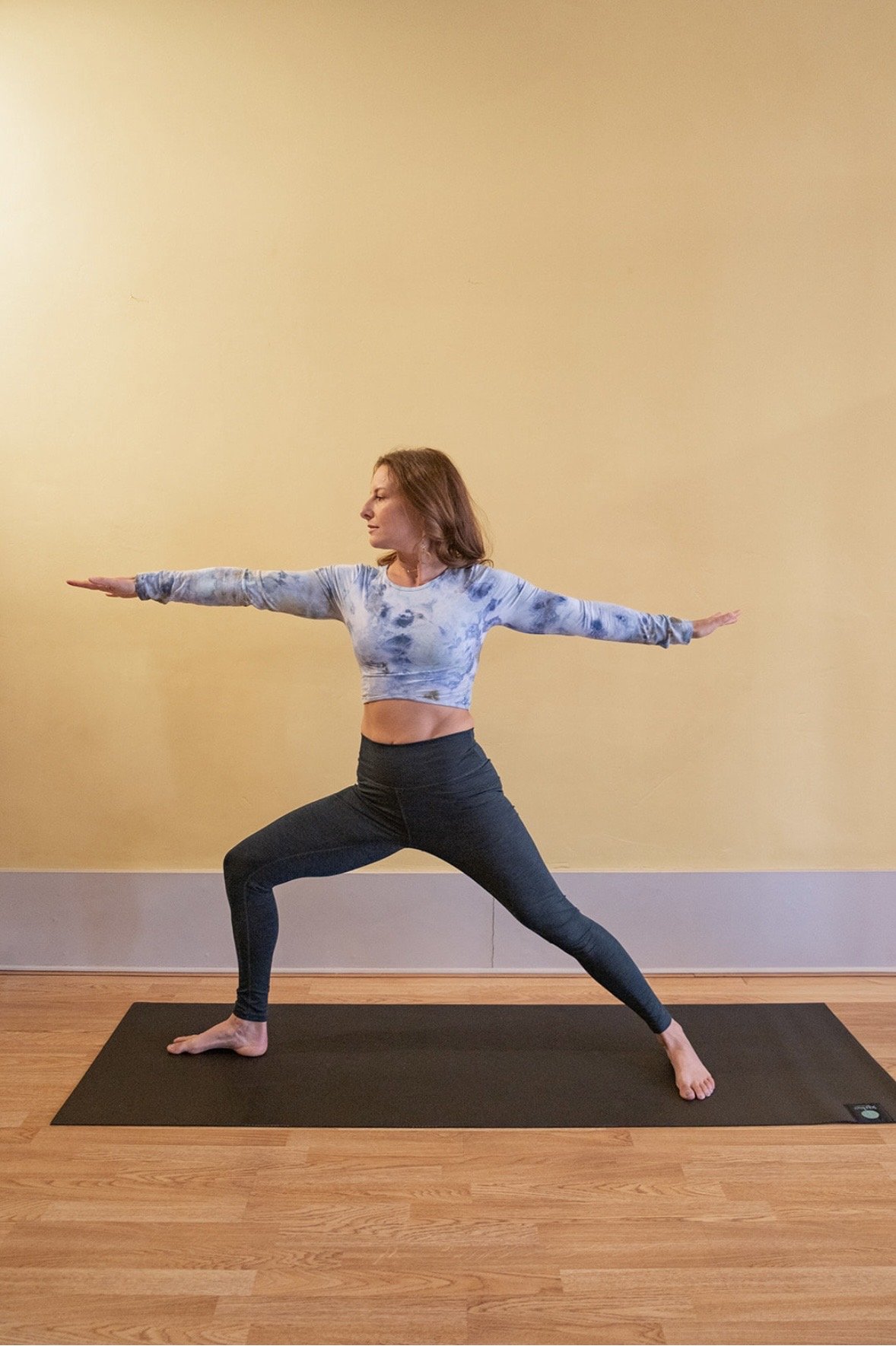 A woman practicing yoga indoors, standing on a black yoga mat with her arms extended sideways and legs in a wide stance, against a plain beige wall.