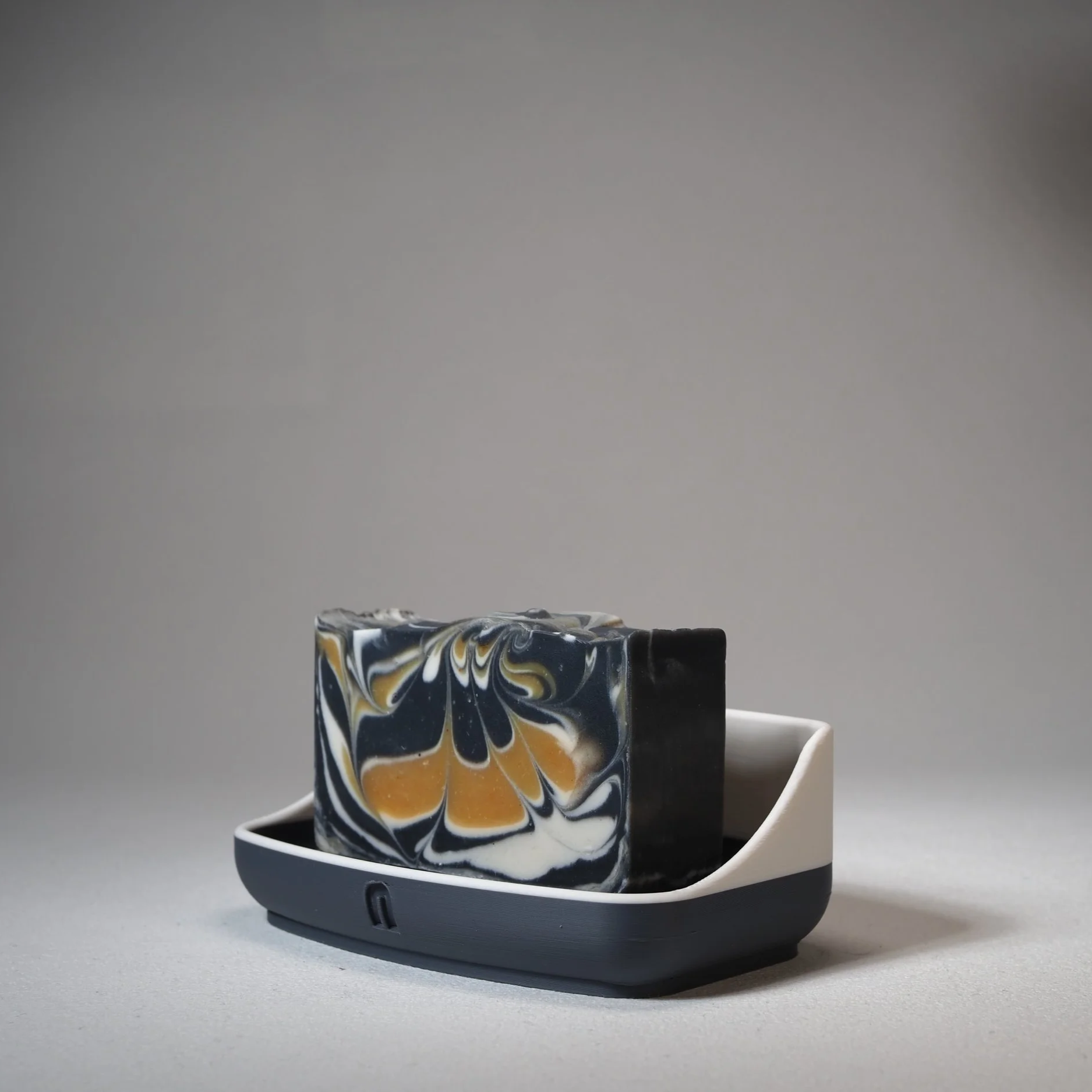 Close-up of a marble-patterned soap bar with black, white, grey, and yellow swirls, resting on a white dish with a robotic vacuum cleaner visible at the bottom. The background is plain and grey.