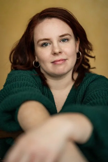 Ruth, a white woman with shoulder-length, slightly burgundy hair and freckles, stands in a playful, leaning-forward pose, smiling broadly. She wears a light blue zip-neck top. The background is a mottled blue-green studio wall.