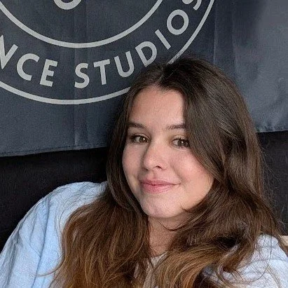 A white woman with brown hair. She is wearing a light blue linen top. She sits behind a dark blue flag with the logo of Valentine Studios.