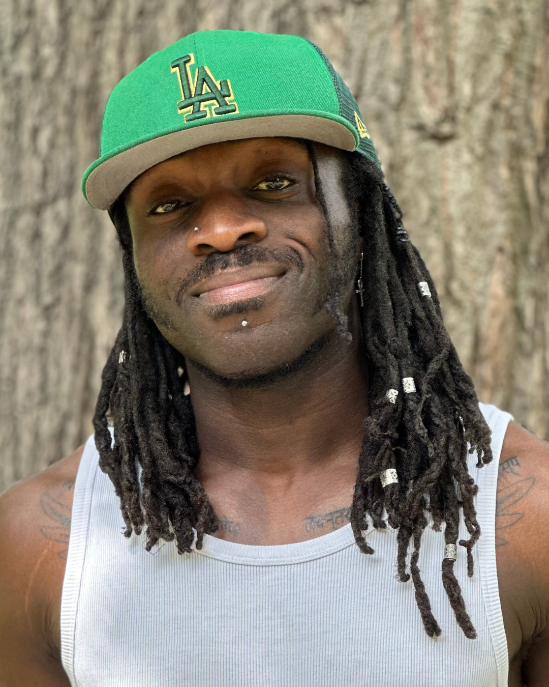 Tiger, a black man with  shoulder length dreadlocks. He poses with a smile. He wears a white vest top and green hat. He has 2 facial piercings on his nose and below his lip. His dreadlocks are decorated wtih silver beads.
