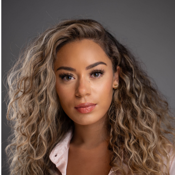 Headshot of a woman with long highlighted curly hair and natural makeup against a grey background.
