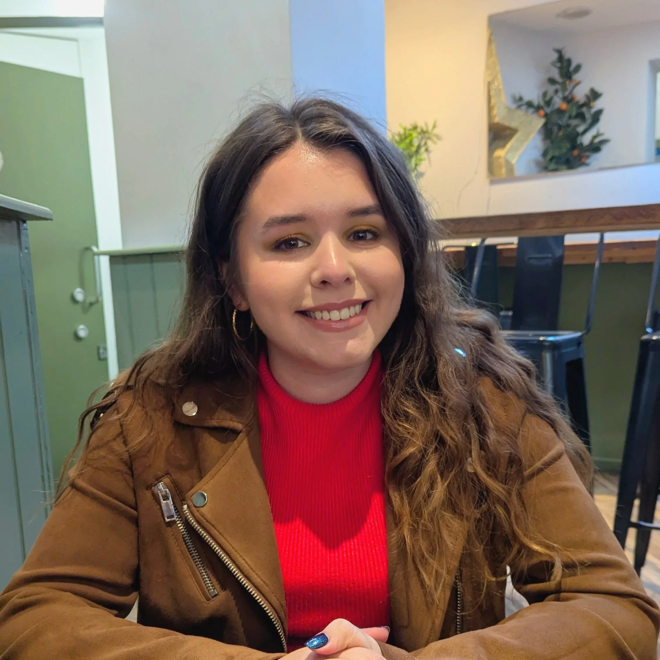 Image of Ines, the founder of Valentine Studios. She is a white woman with brown hair. She is wearing a orange ribbed tank top and suede jacket. She is sitting in a cafe.