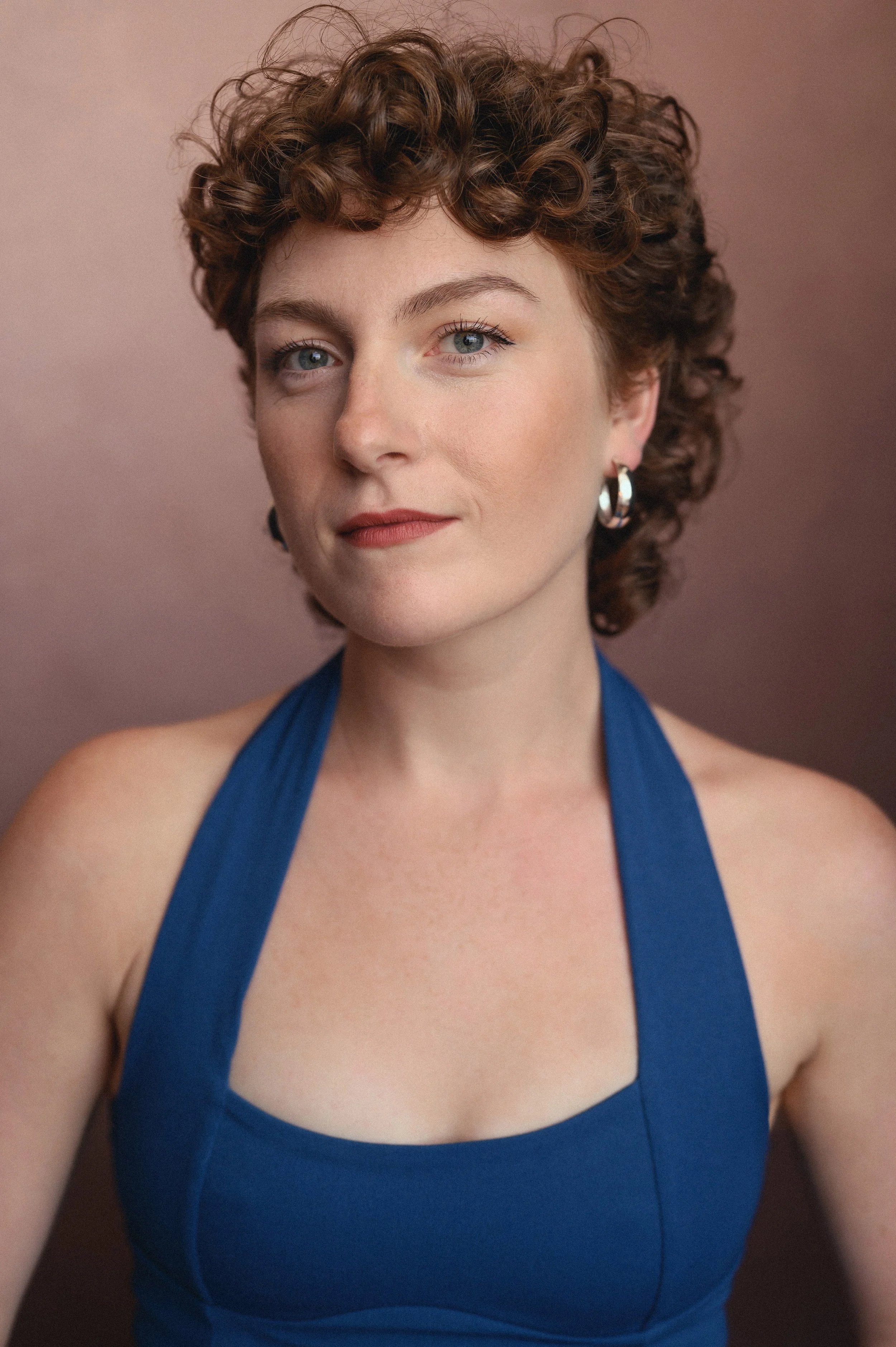Portrait of a person with short, curly auburn/brown hair wearing silver hoop earrings and a deep blue halter-style top, looking directly at the camera with a warm, focused expression against a warm brown background.