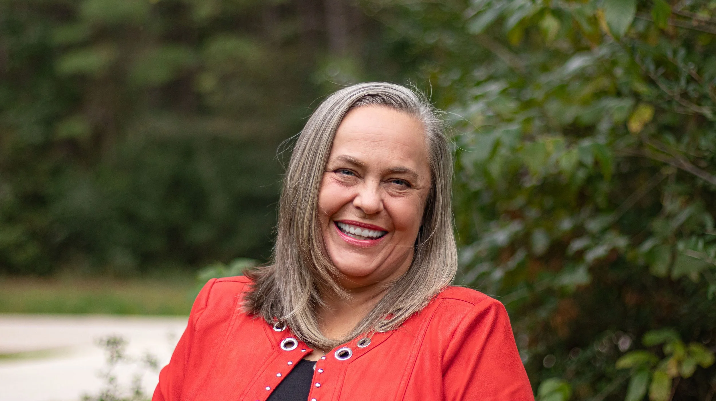 A woman with gray hair smiling outdoors, wearing a red jacket and a black top, standing in front of green foliage.
