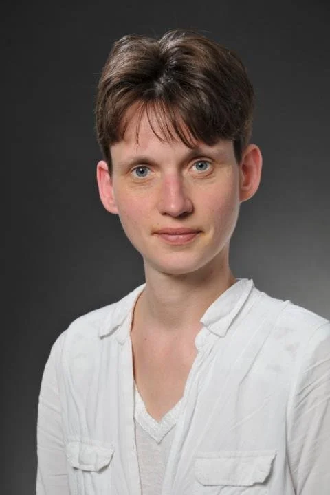 Portrait of a woman with short brown hair, blue eyes, wearing a white shirt against a dark gray background.