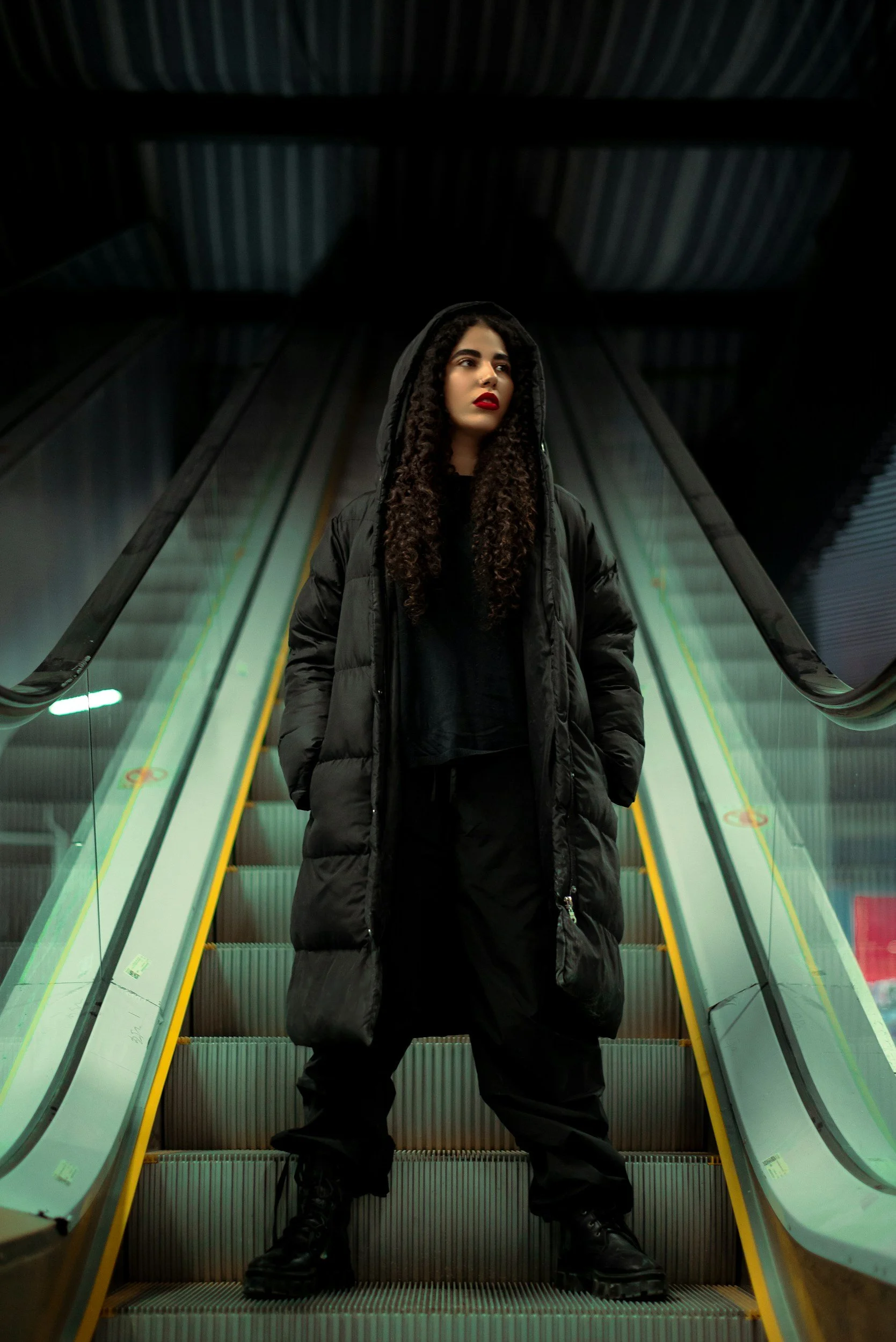 A streetwear style fashion image where the model wears a black hooded puffer jacket and striking red lipstick. The model is against an underground escalator.