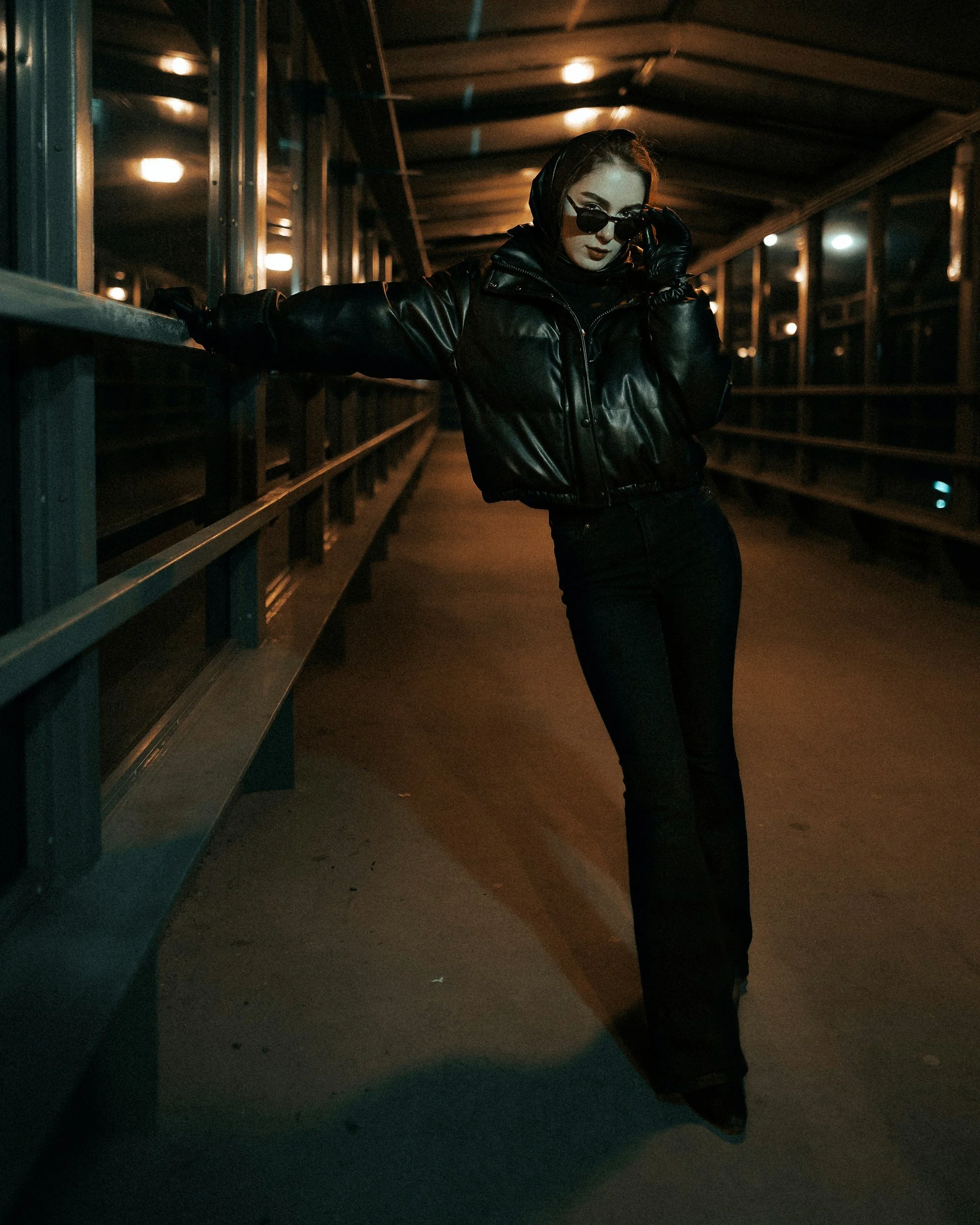 A cinematic fashion image of a model wearing all black peering over the top of black sunglasses. She is photographed stood in a sheltered overpass with teal and orange lighting.