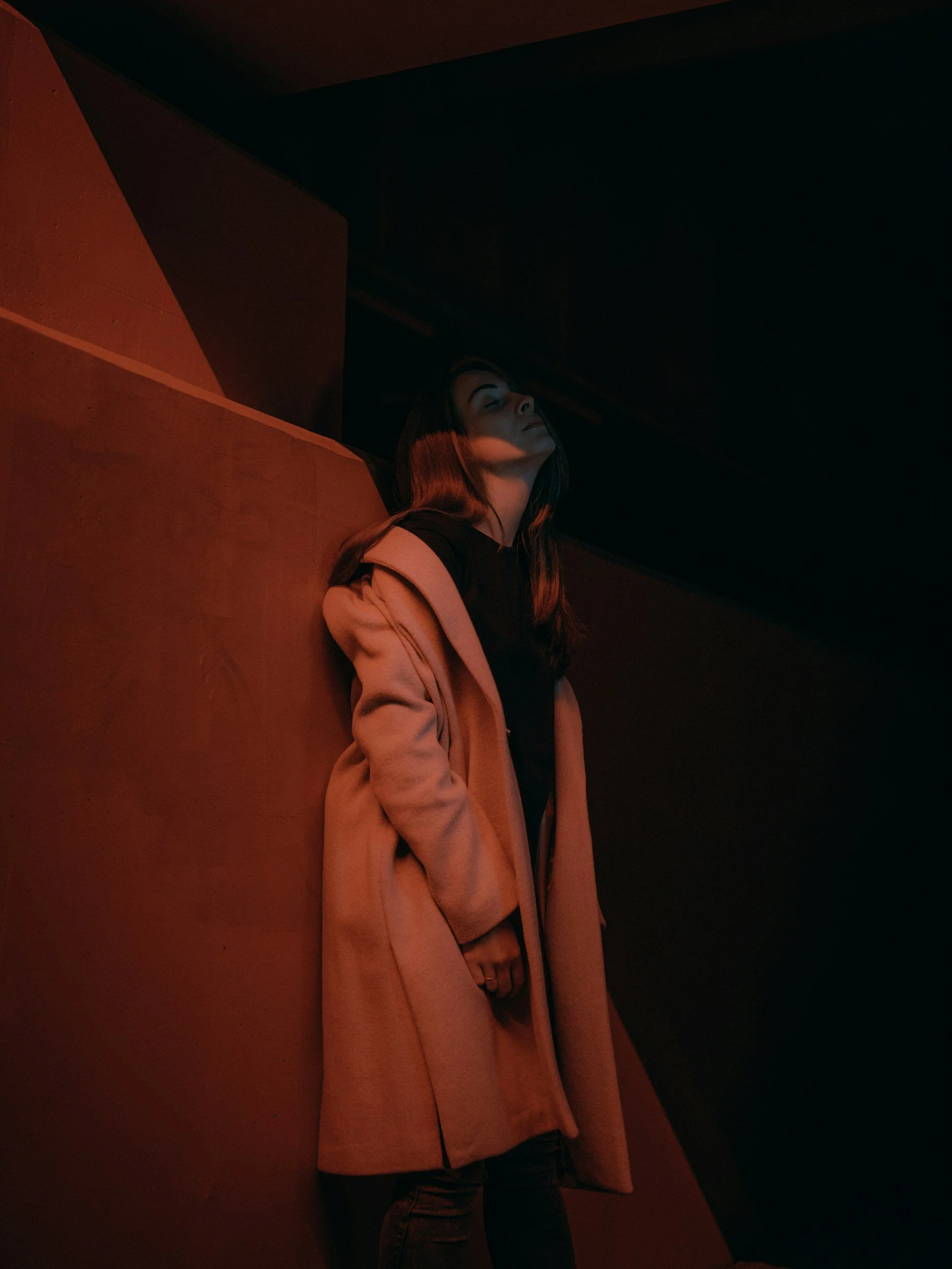 A woman with long hair wearing a beige coat and black shirt standing against a red wall with dark background.