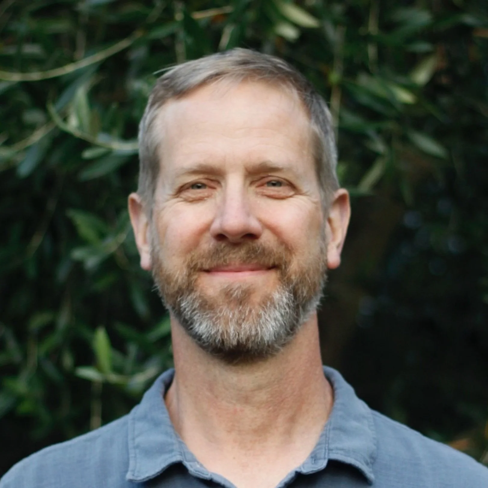 A middle-aged man with a beard and short hair, smiling, standing outdoors with green foliage in the background.
