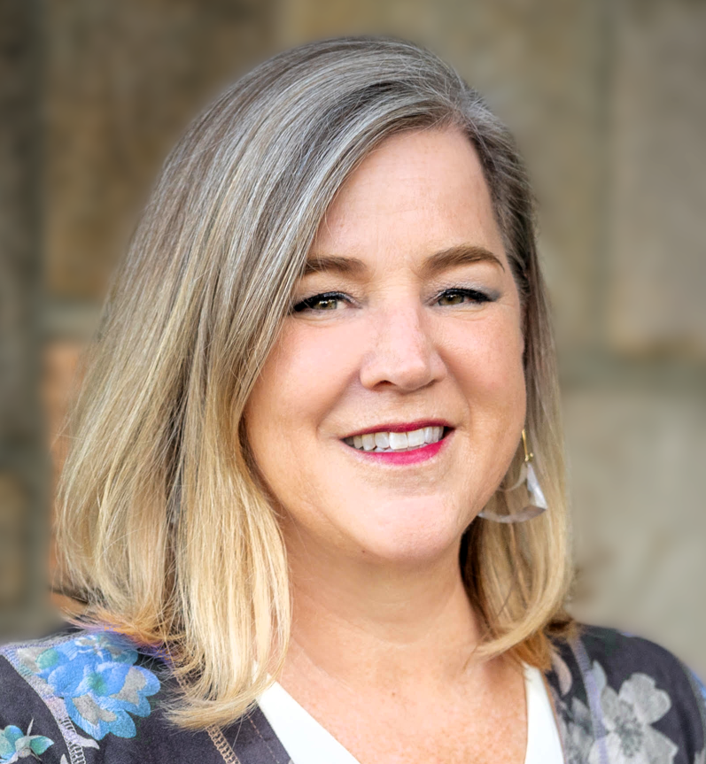 A smiling woman with blonde hair and blue eyes, wearing a floral blazer and a white top, standing outdoors with a blurred stone wall background.