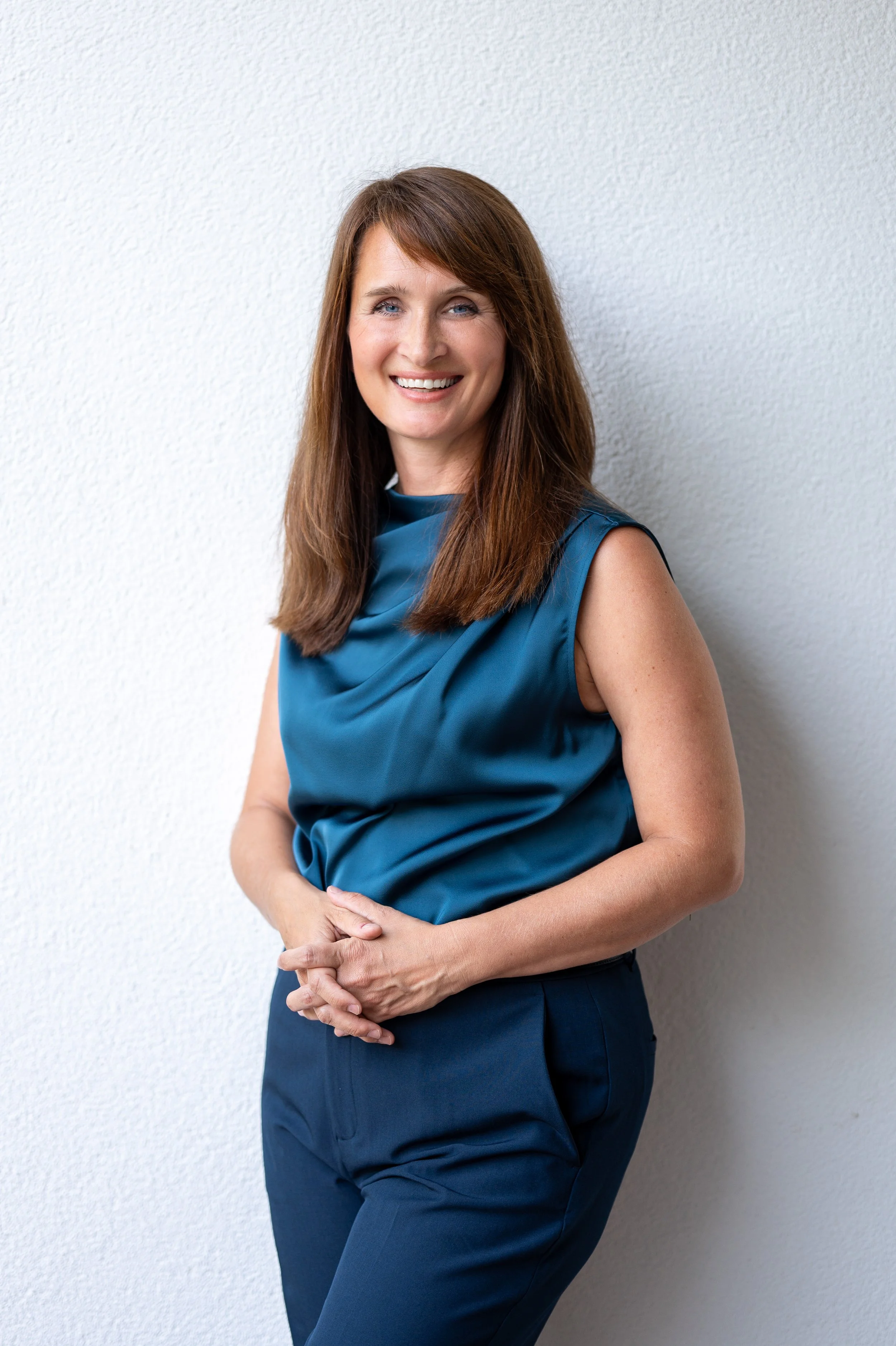 A woman with long, wavy reddish-brown hair smiling outdoors in front of a stone wall, wearing a white blazer and coral blouse.